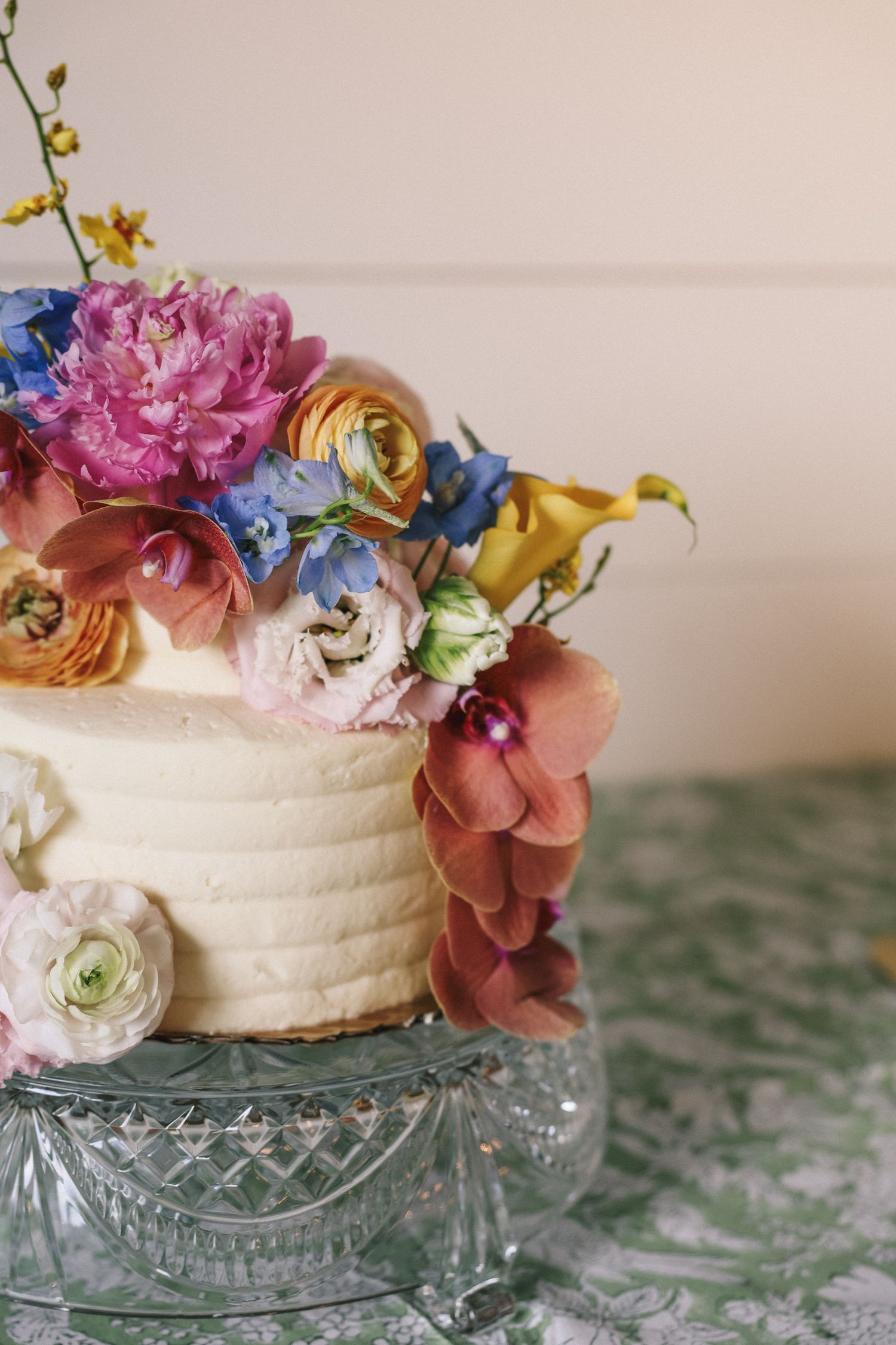 White wedding cake decorated with pink, blue and orange fresh flowers on a crystal cake stand.