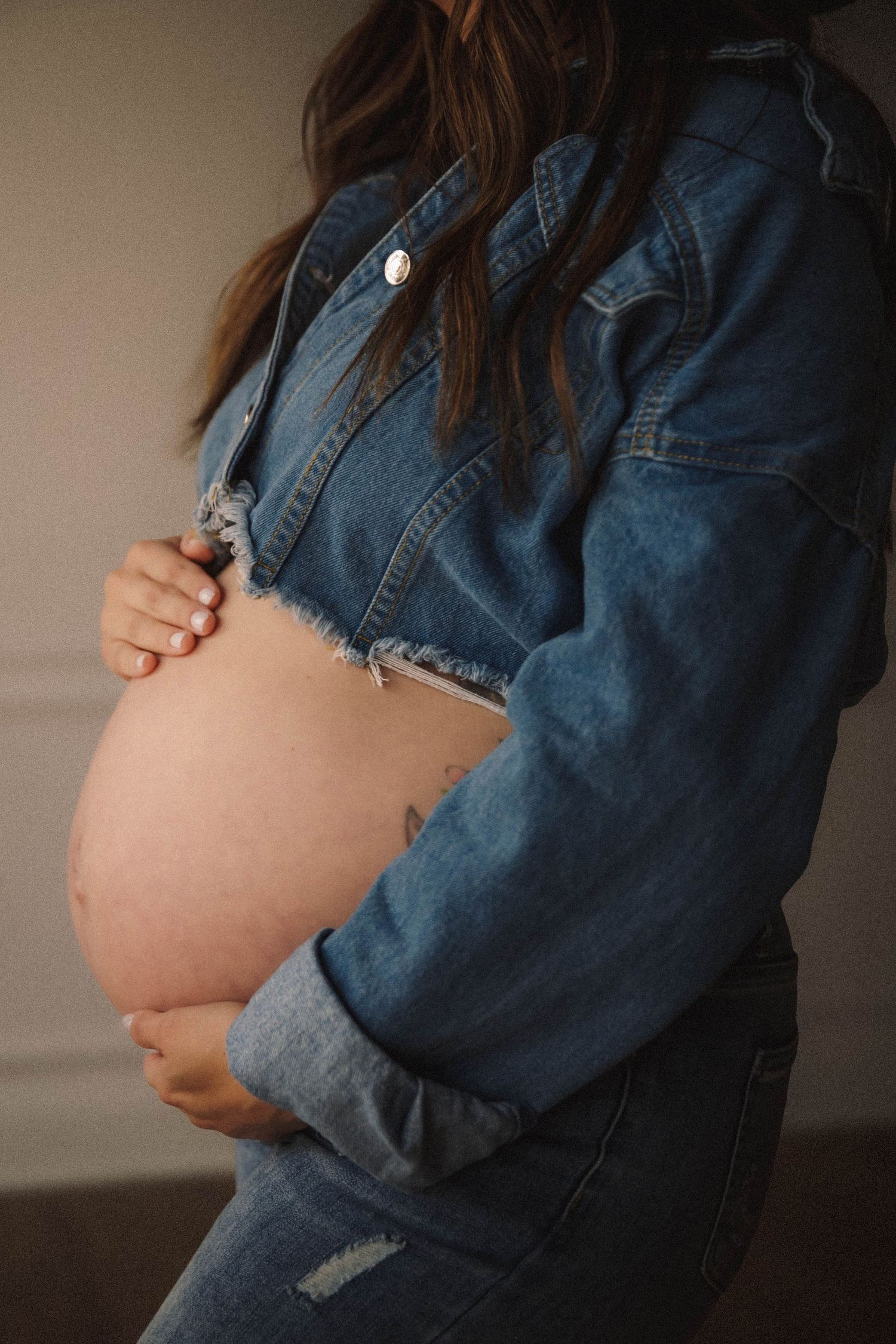 Side view of pregnant belly with person wearing denim jacket and dark pants, photographed against neutral background.