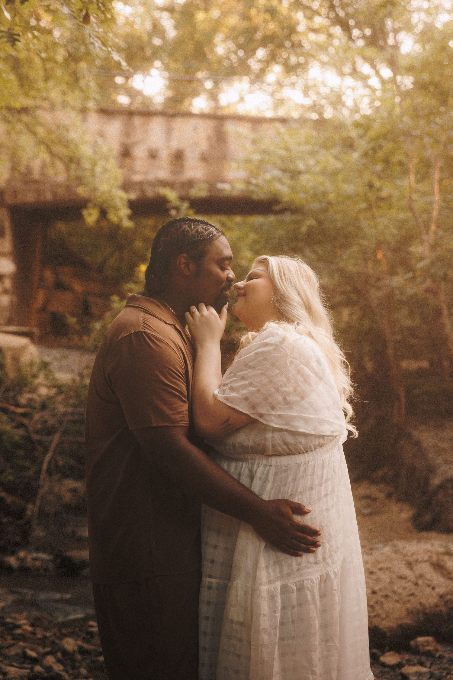 A romantic moment captured outdoors with a rustic bridge in the background during golden hour.