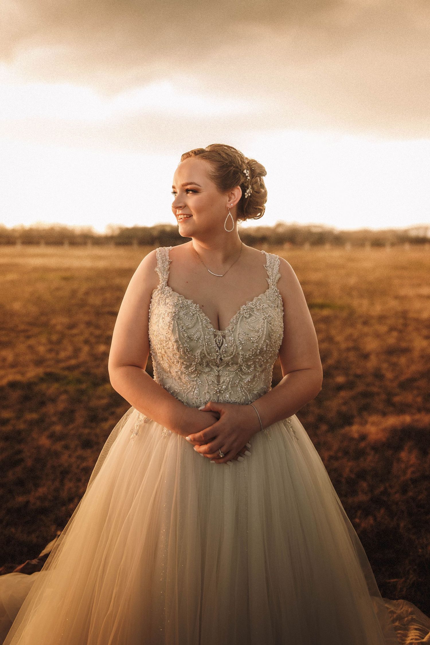 A bride wearing a stunning embellished ballgown poses in a golden field at sunset for romantic wedding portraits.