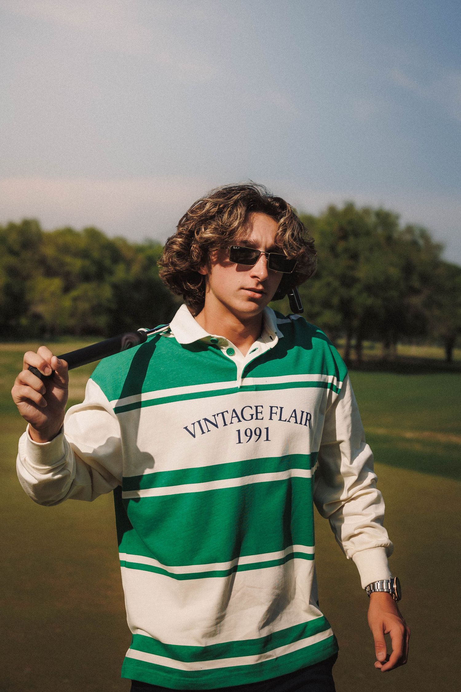Person in green and white striped vintage rugby shirt poses outdoors on sunny field with trees in background.