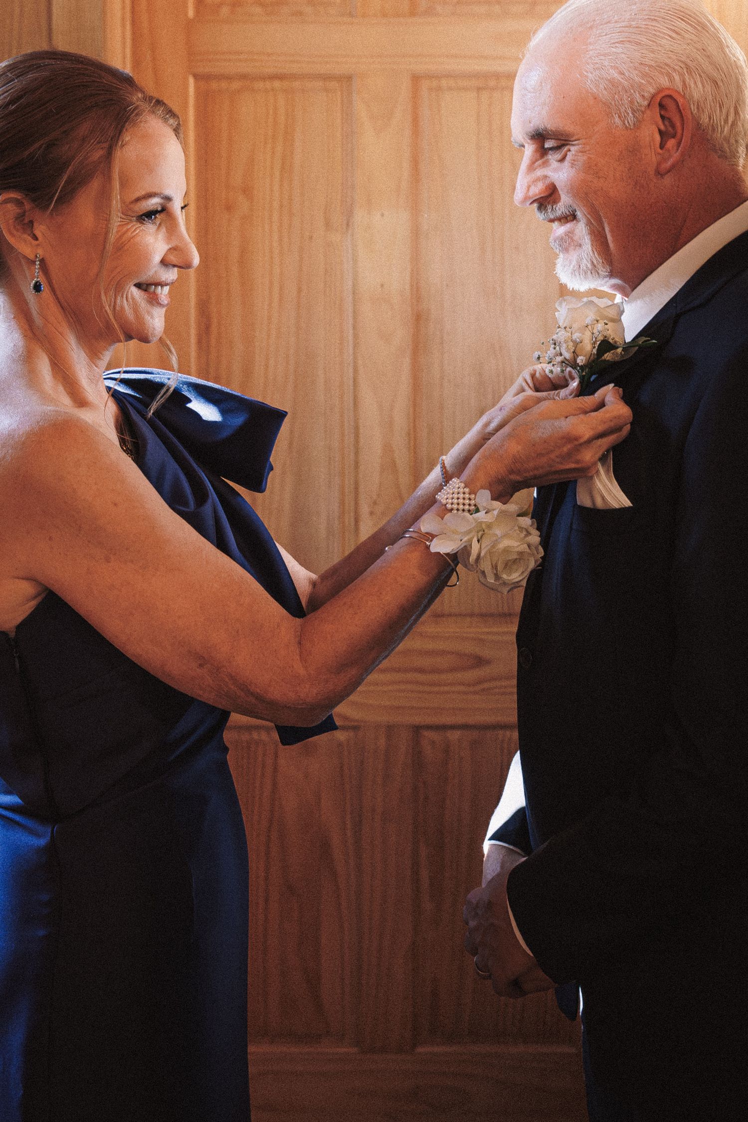 Woman in blue gown pins boutonniere on older man in black suit at wedding.