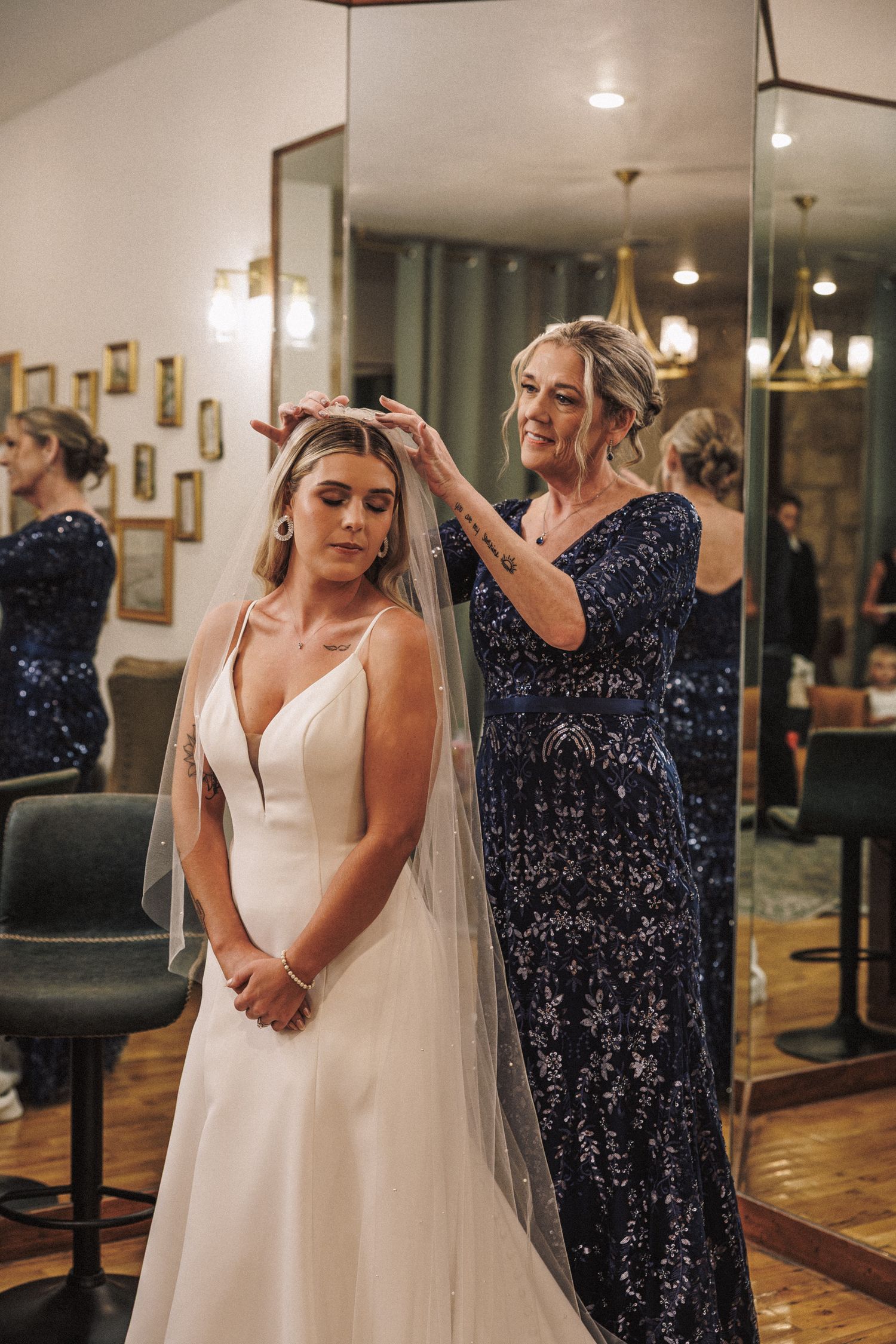 Mother in navy sequin gown placing veil on bride in elegant white wedding dress before the ceremony.