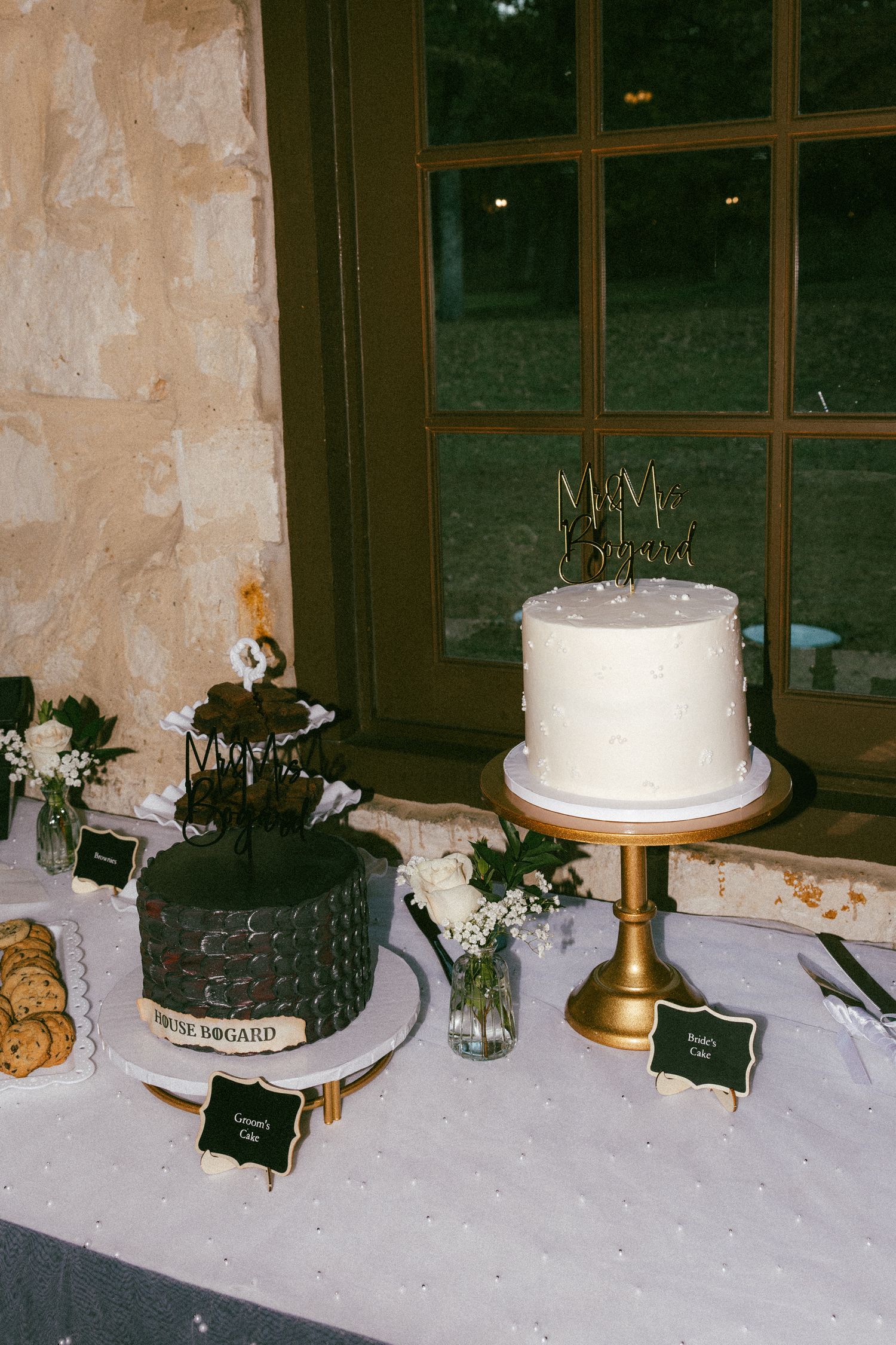 Elegant two-tier white wedding cake beside dark grooms cake on decorated dessert table.