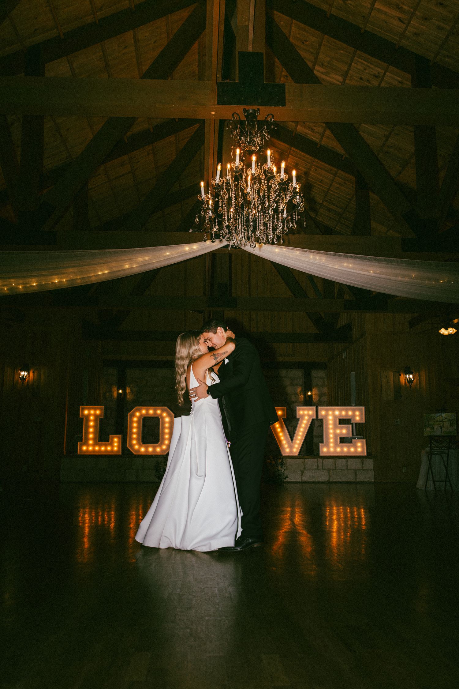 Bride and groom share first dance beneath chandelier with illuminated LOVE marquee letters in barn.