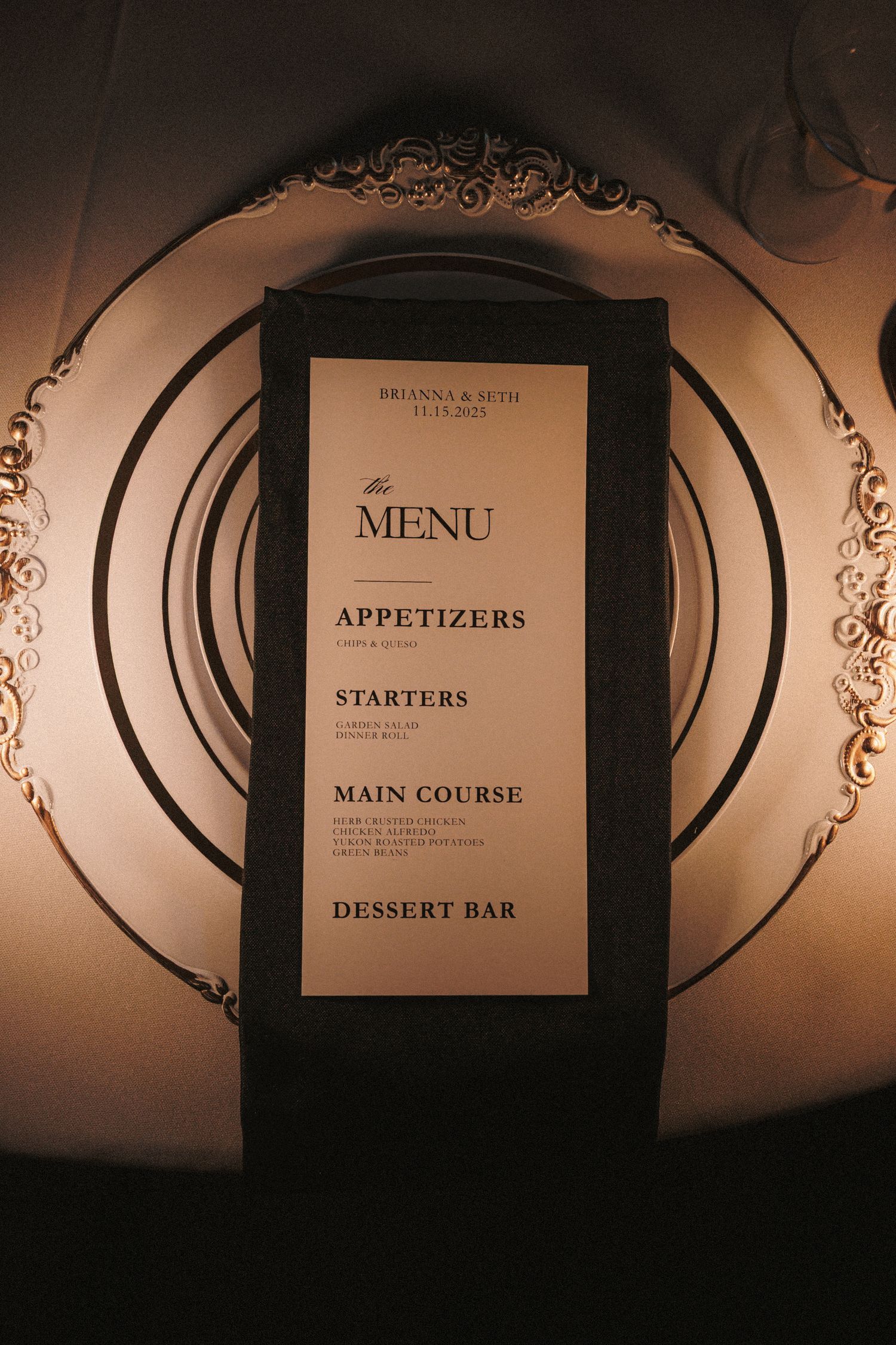 Elegant wedding menu card on black napkin atop decorative gold charger plate featuring appetizers, starters, main course, and dessert bar.