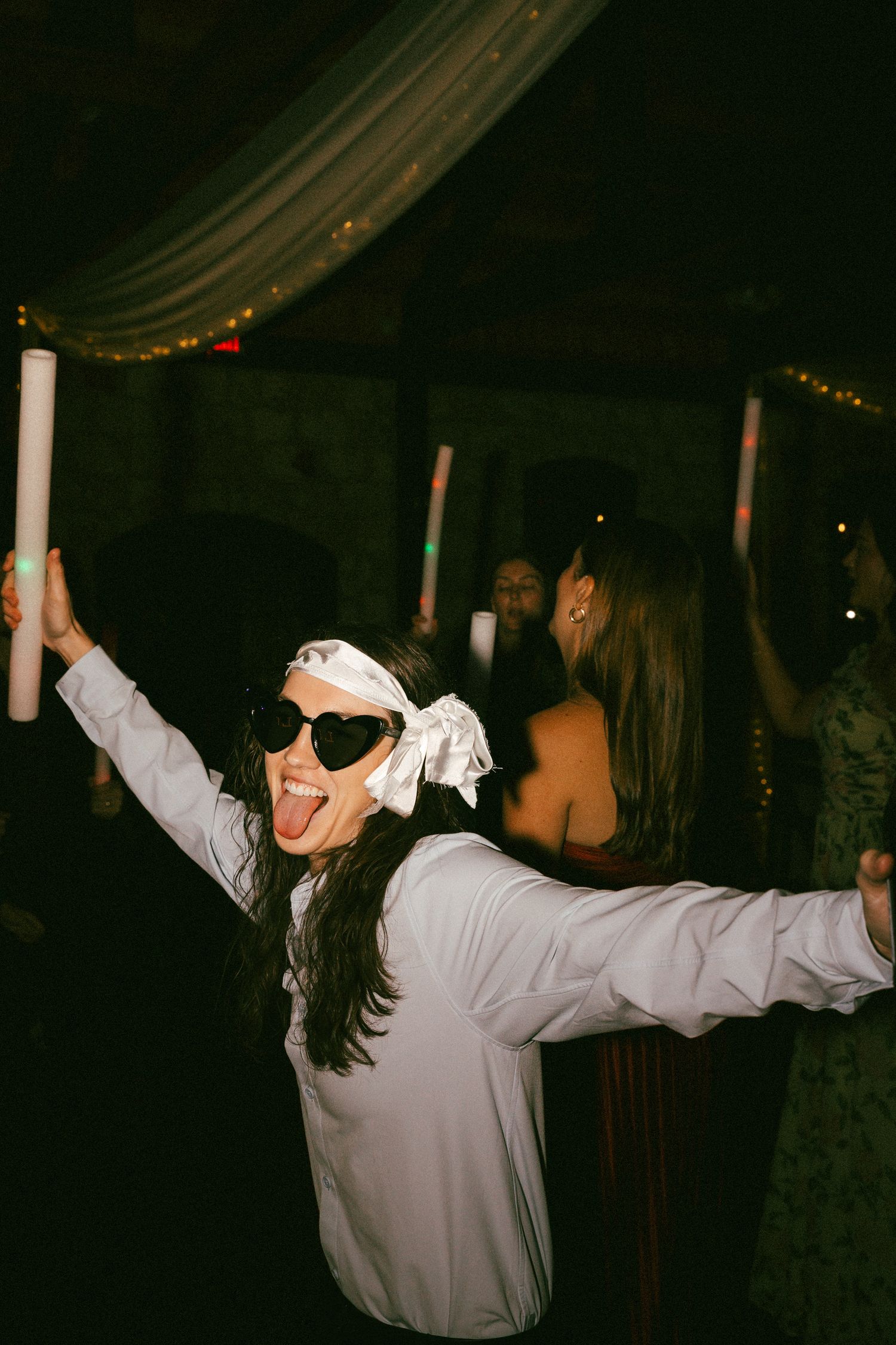 A joyful woman in heart sunglasses and white shirt dances with a glow stick at a wedding reception.