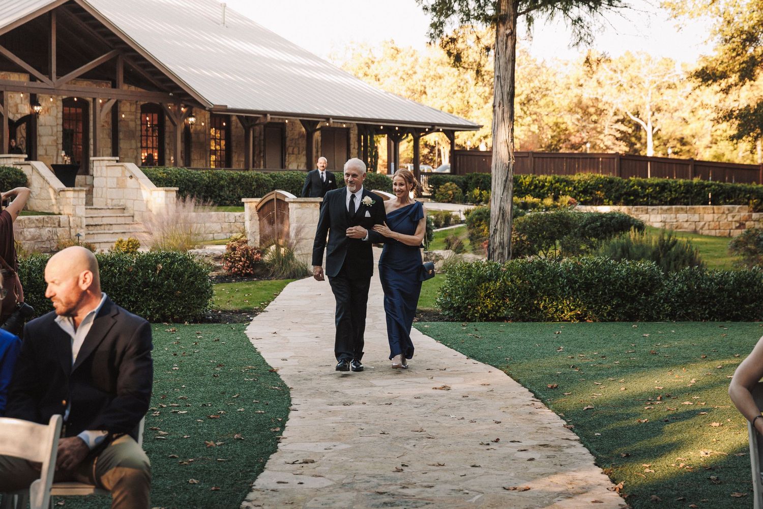 Couple walks down outdoor wedding aisle toward rustic venue surrounded by lush gardens and autumn trees.
