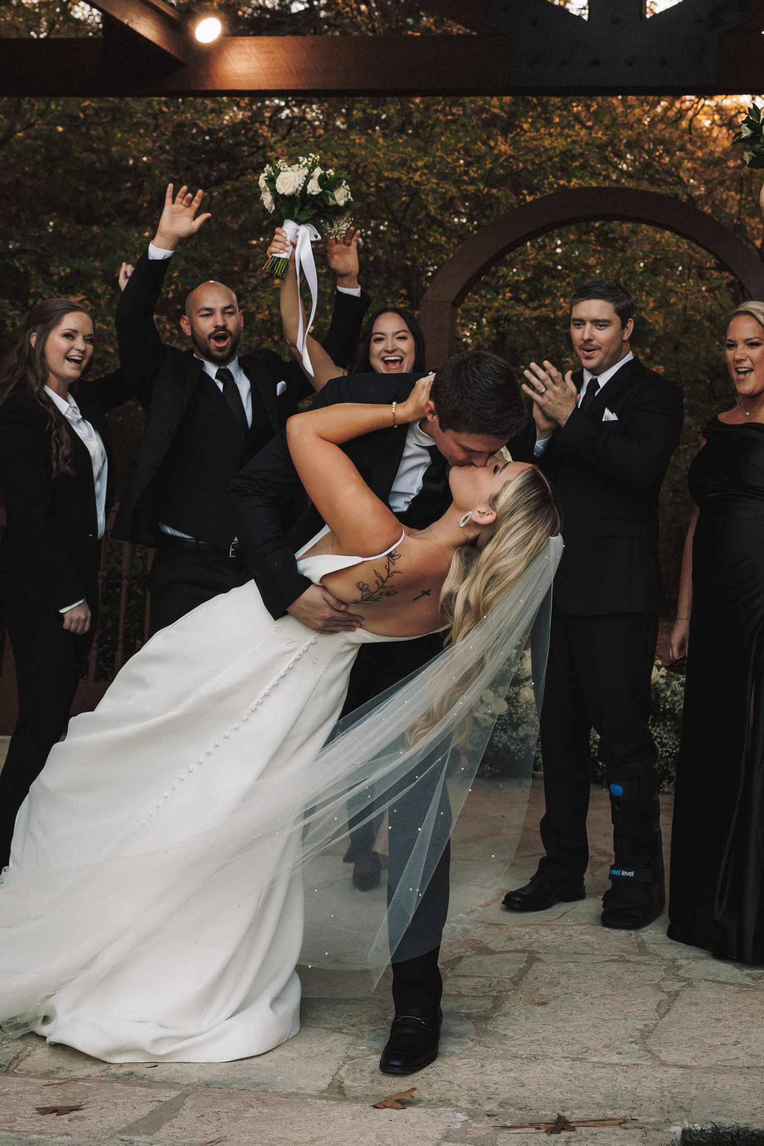 Groom dips bride for romantic kiss surrounded by cheering wedding party outdoors under rustic arbor.