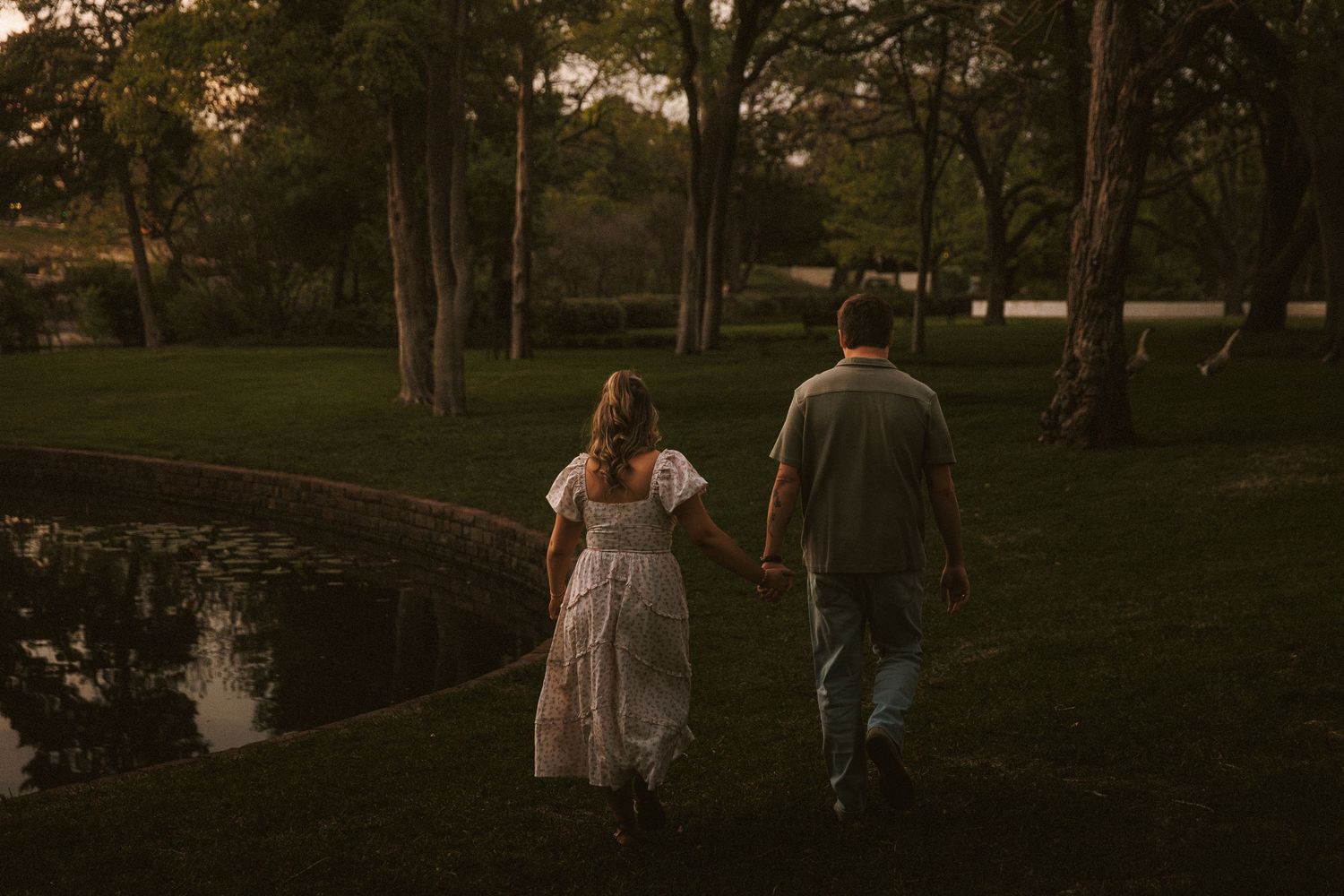 A couple walks hand-in-hand through a lush green park beside a peaceful pond at dusk.