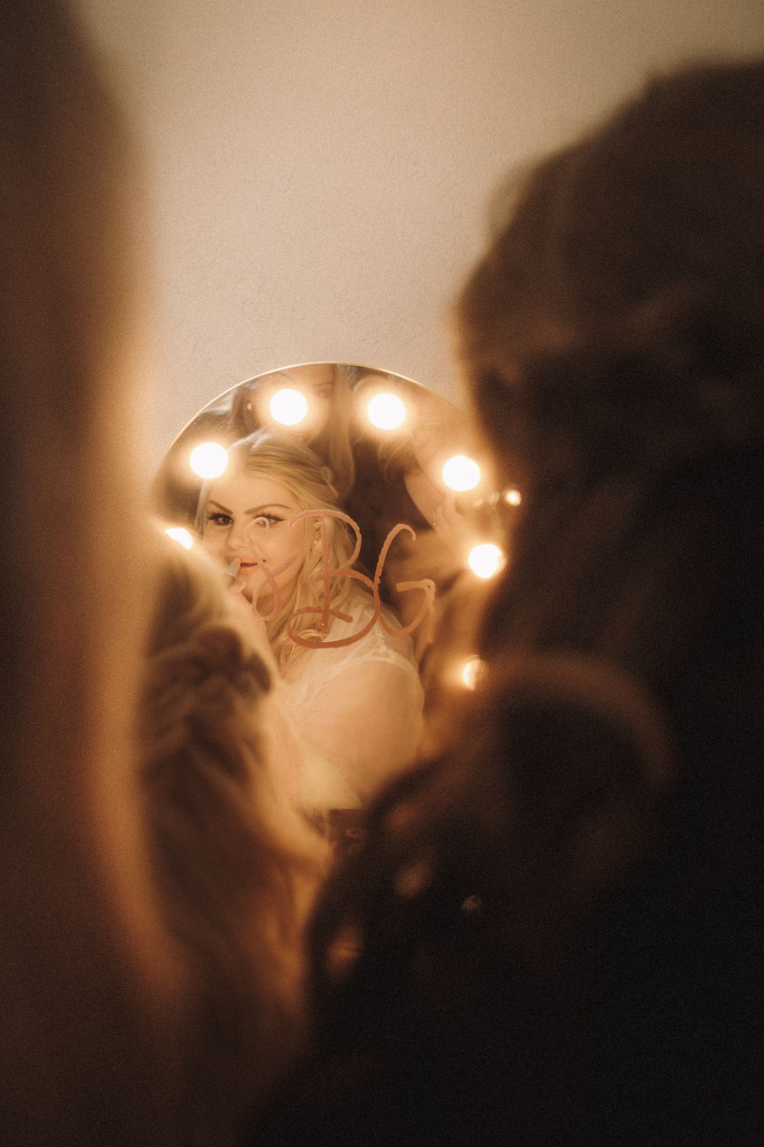 Bride smiling and sticking out tongue reflected in a round Hollywood bulb vanity mirror.