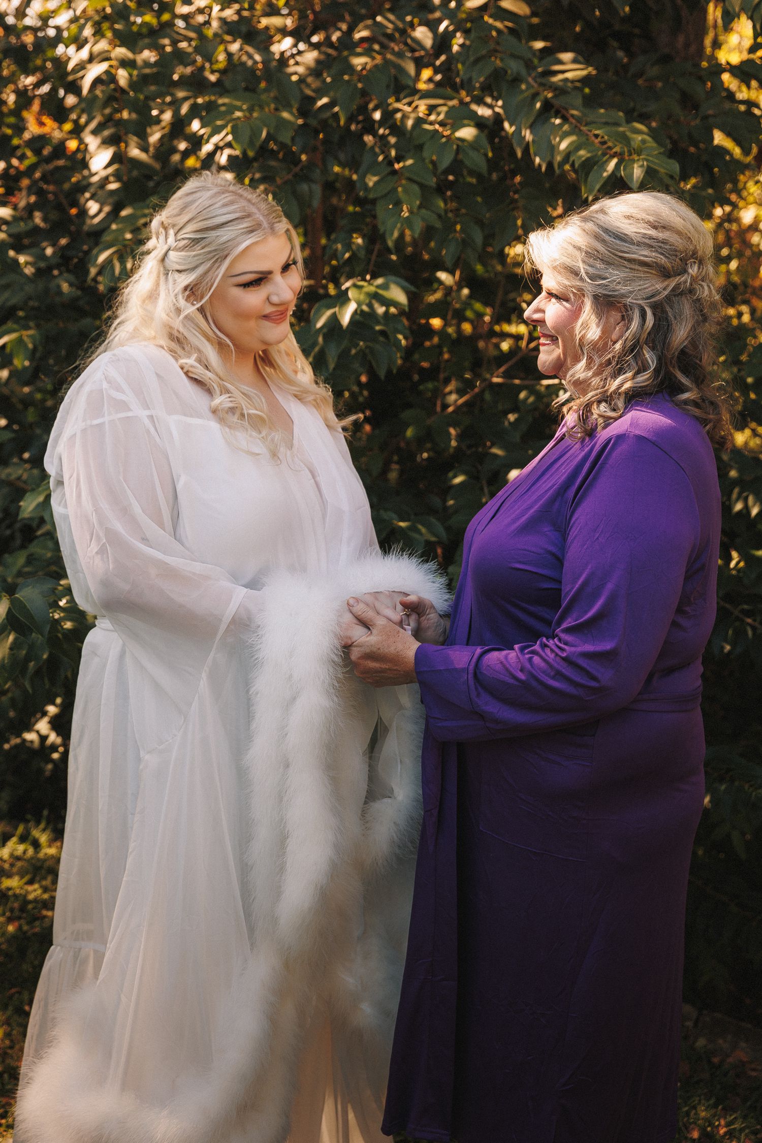 Bride in white tulle gown and mother in purple dress share a tender moment outdoors at golden hour.