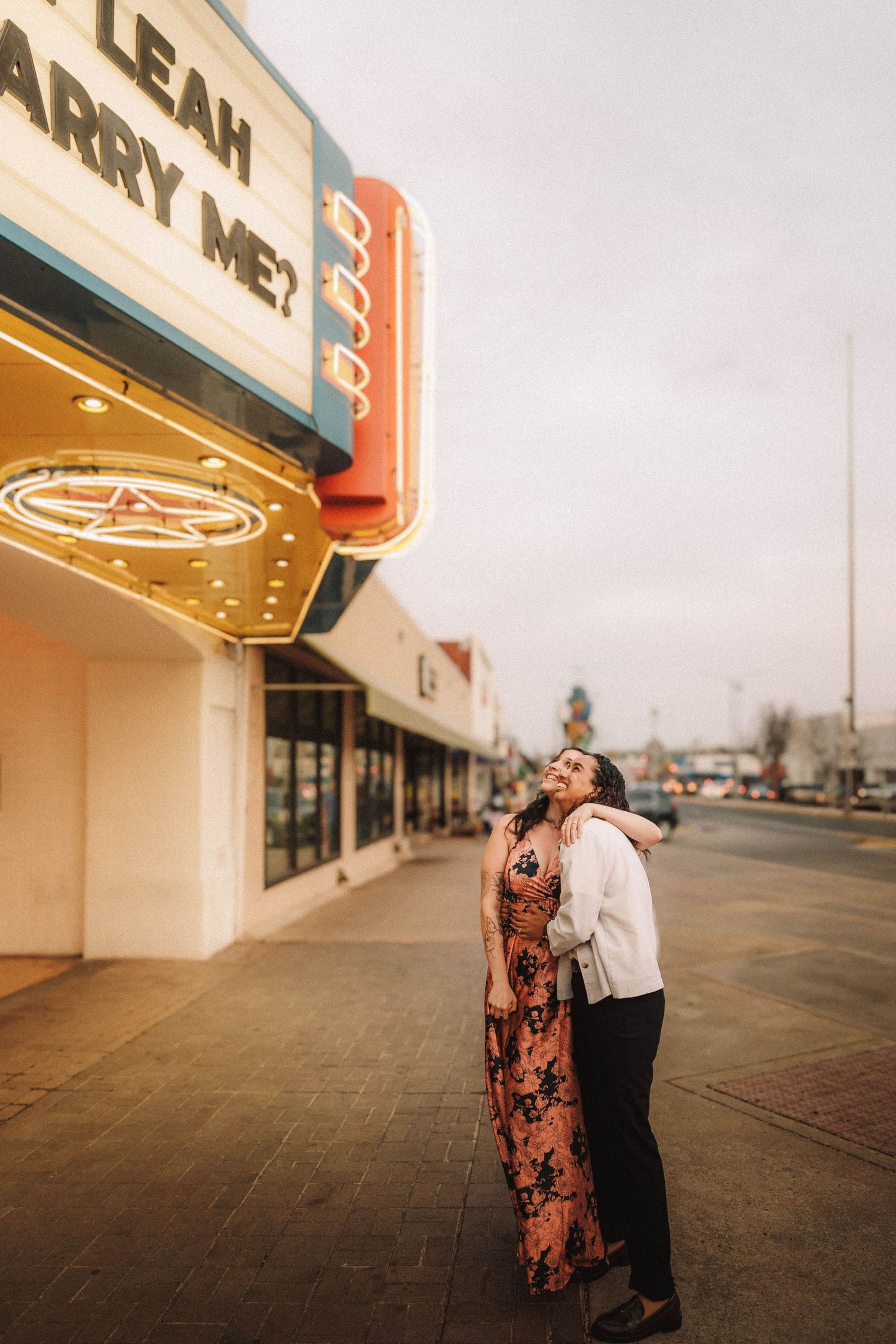 Couple embraces under a theater marquee reading My Lovely Leah Will You Marry Me.
