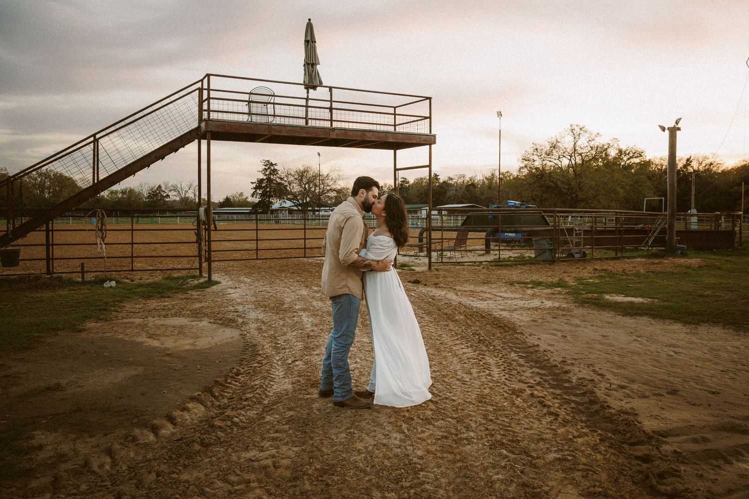 Couple embracing on a dirt path near a rodeo arena during a warm sunset.