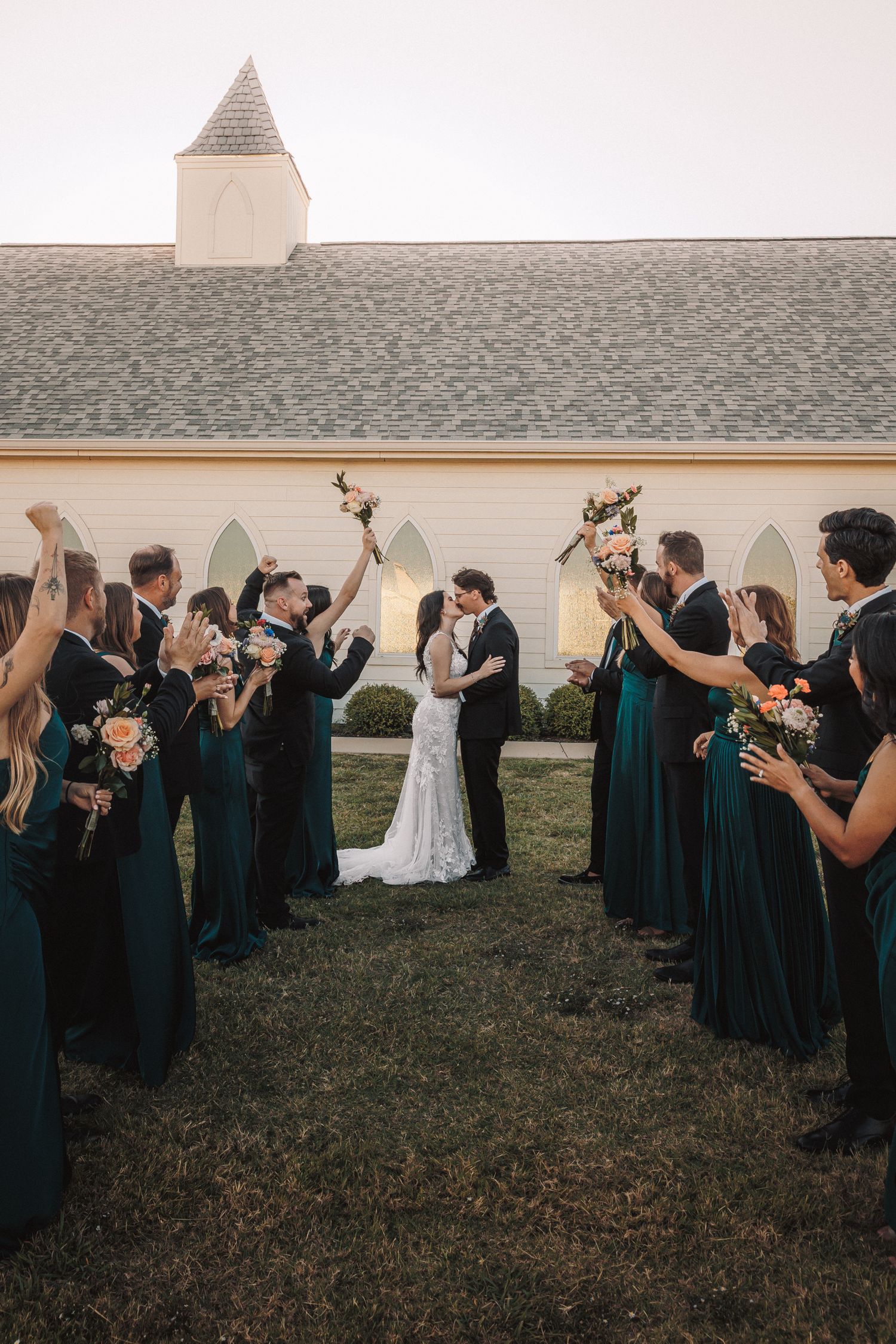 Bride and groom kiss between cheering wedding party in teal gowns outside a white chapel at dusk.