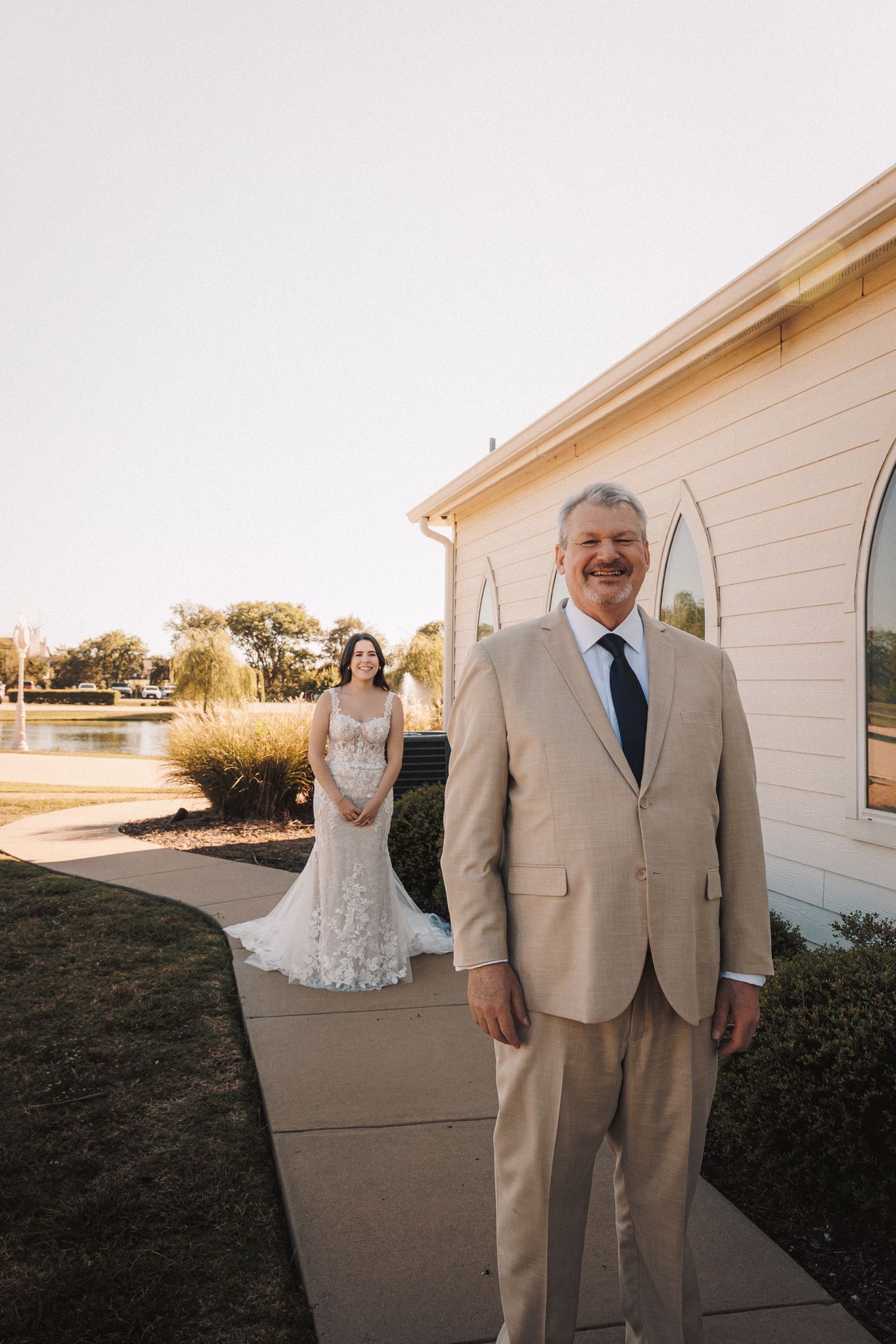 Father of the bride smiling in tan suit as bride in lace gown stands behind him near white chapel.
