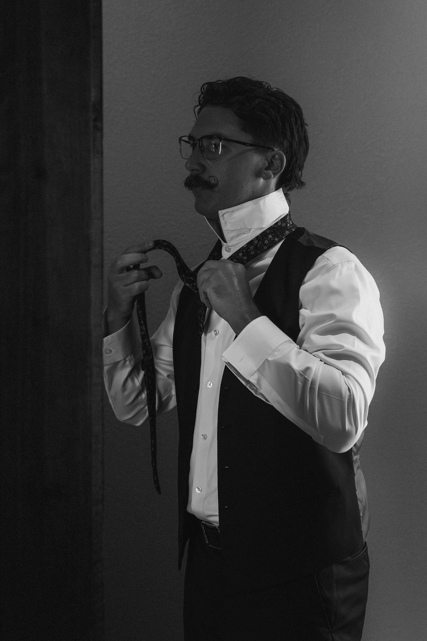 Black and white portrait of a groom adjusting his tie while wearing a vest and glasses.