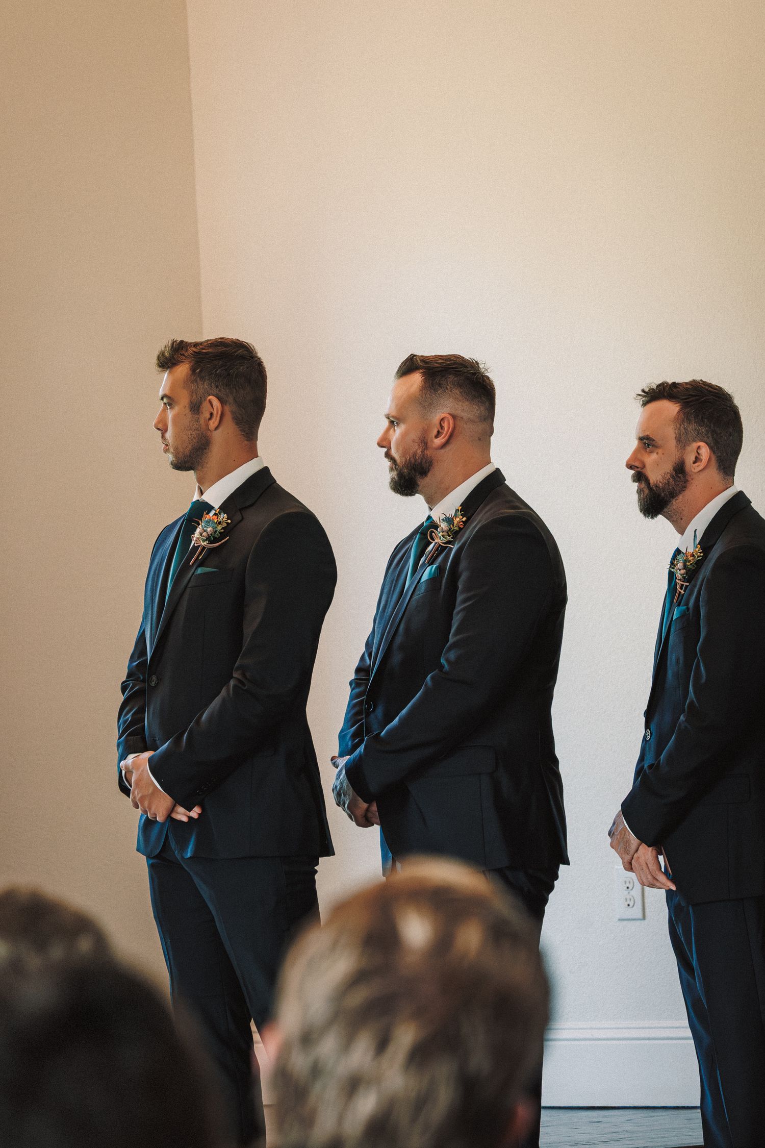 Three groomsmen in black suits with teal ties and boutonnieres stand in a row during a wedding ceremony.