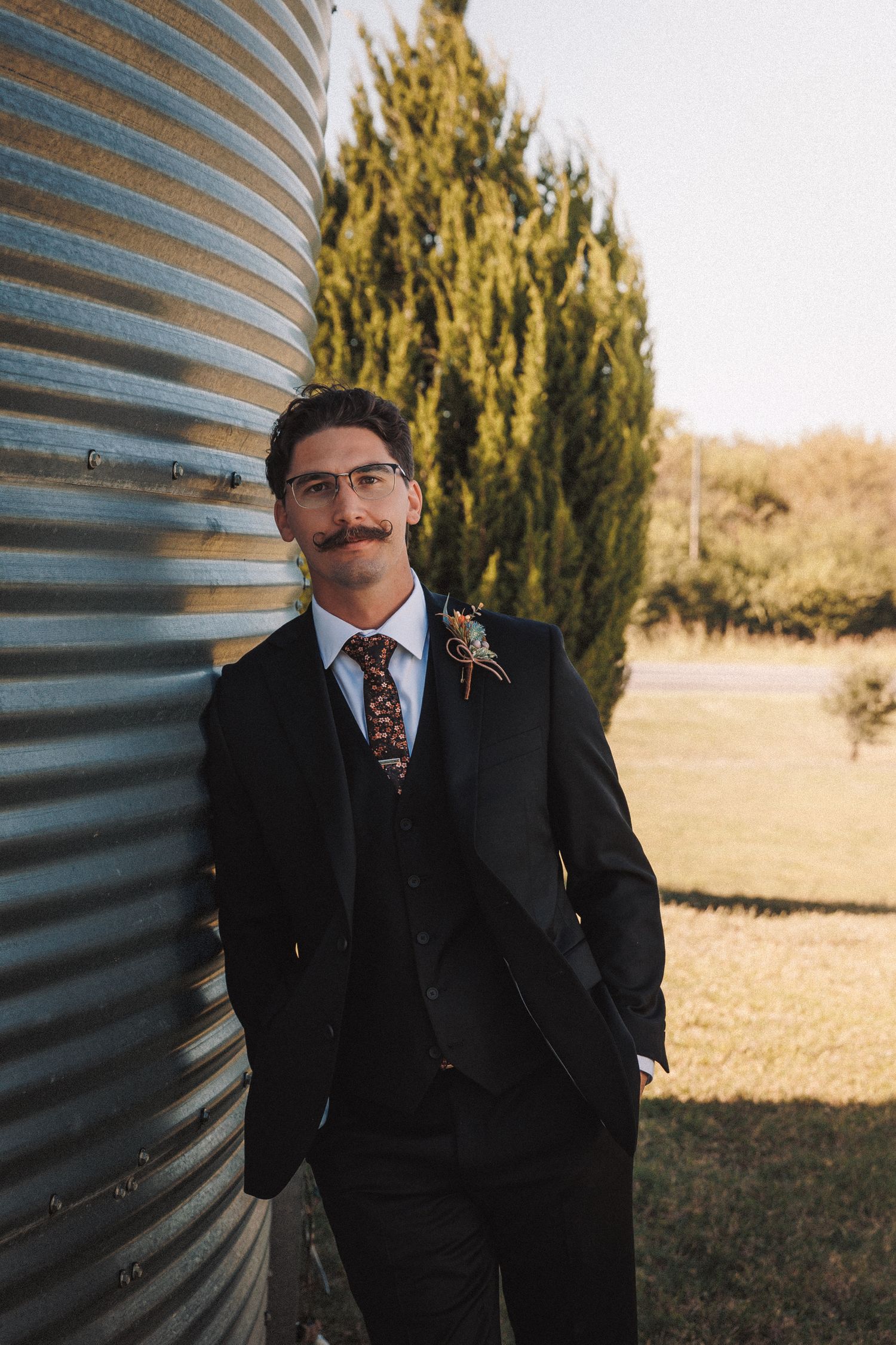 Stylish groom with mustache in black suit and floral tie leans against rustic metal grain silo.