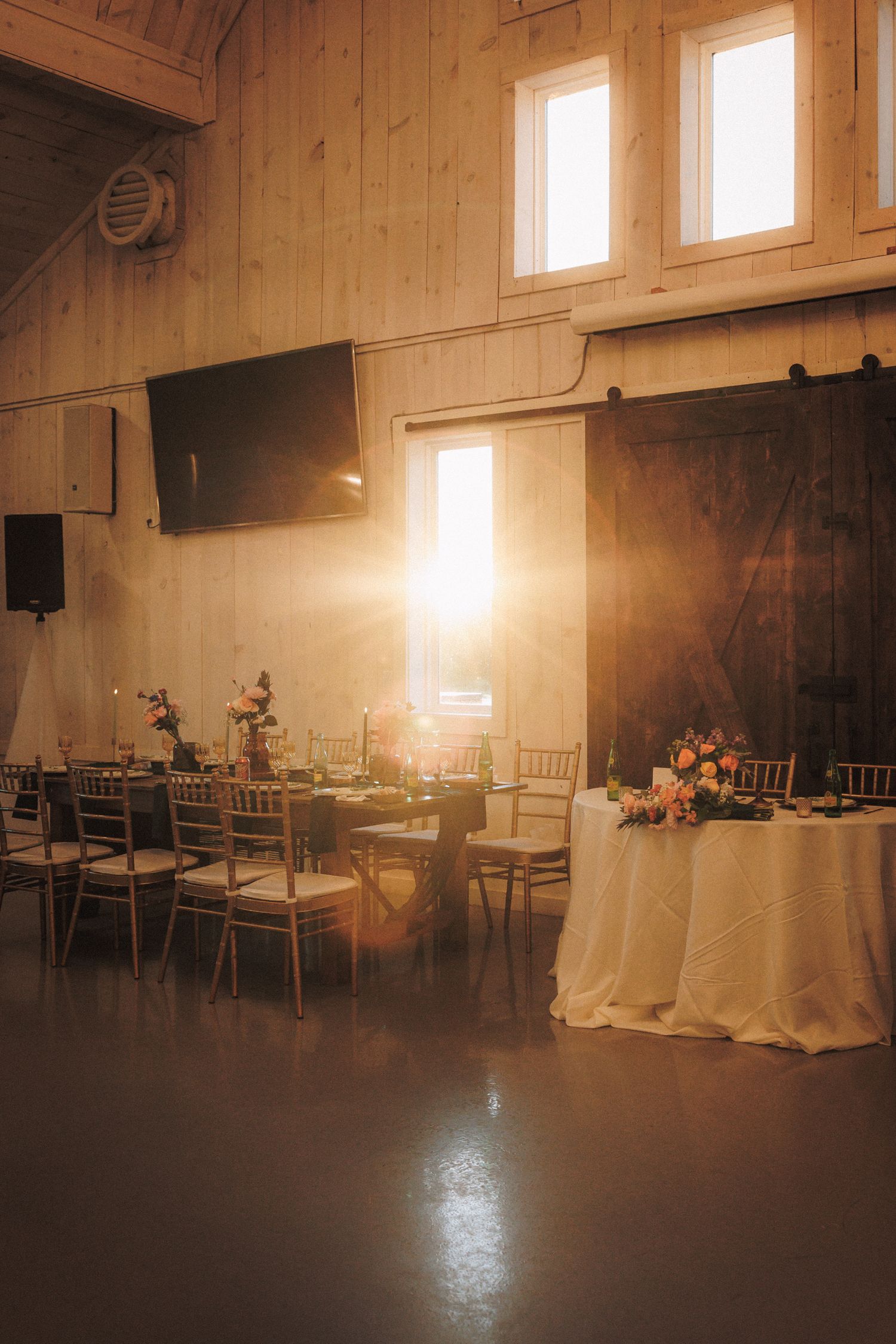 Sunlit barn wedding reception venue with elegant table settings and golden hour light streaming in.