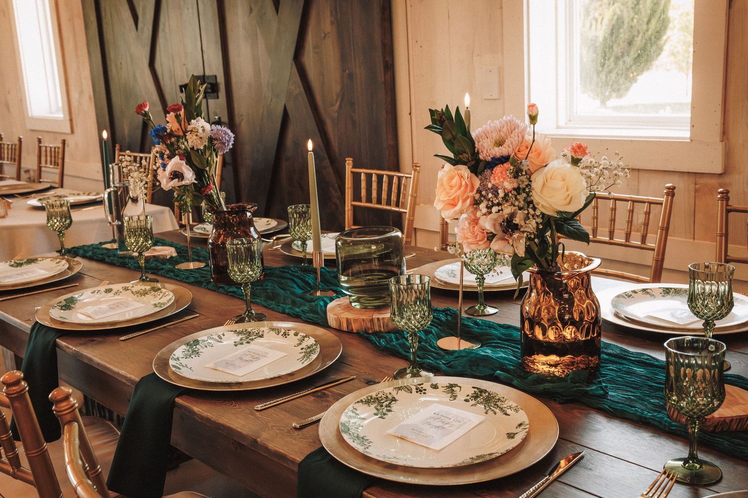 Elegant wedding reception table with teal velvet runner, floral centerpieces, gold cutlery, and green glassware on a wooden table.