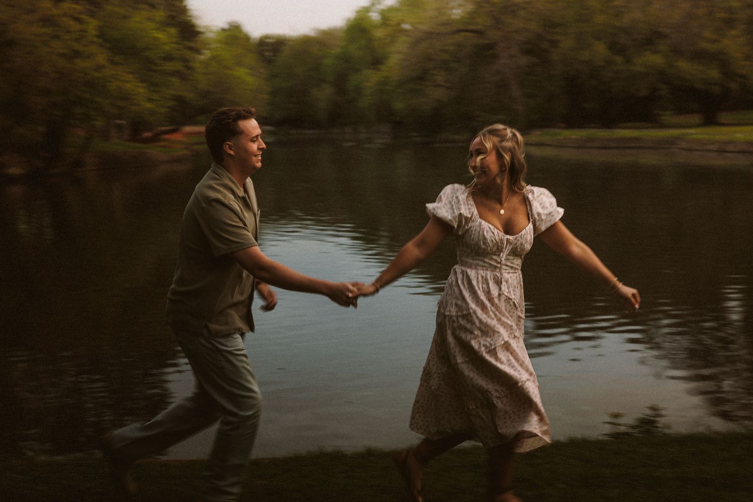 Couple holding hands and running through a serene lake surrounded by lush green trees during a romantic outdoor photo session.