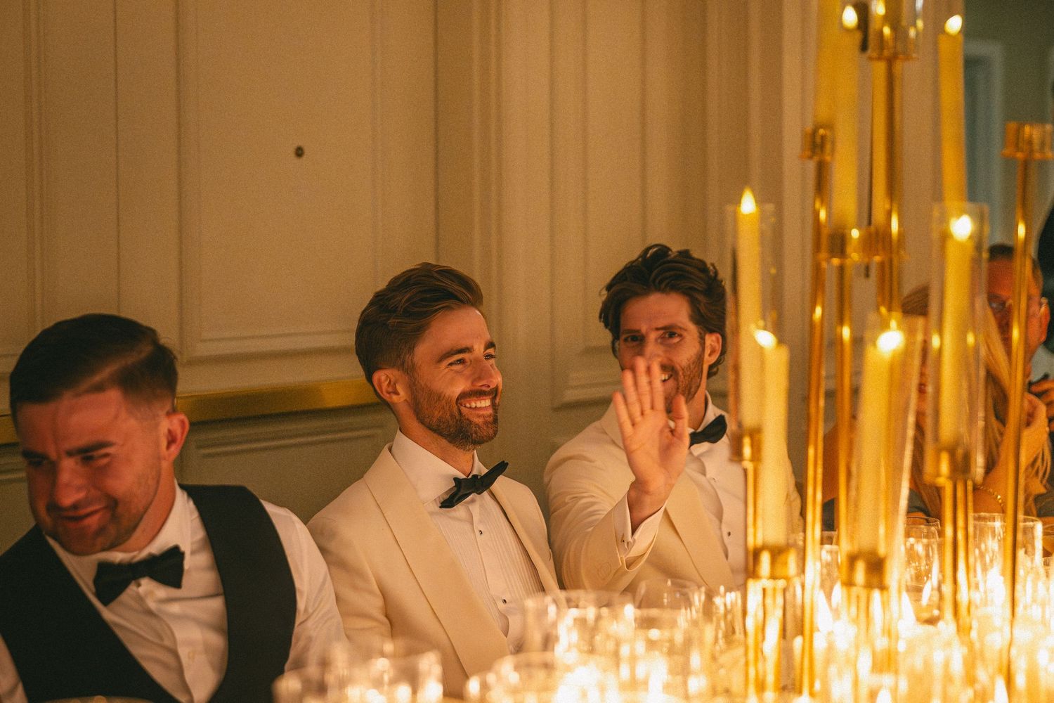 Three men in formal attire laughing at a candlelit wedding reception table with tall gold candelabras.