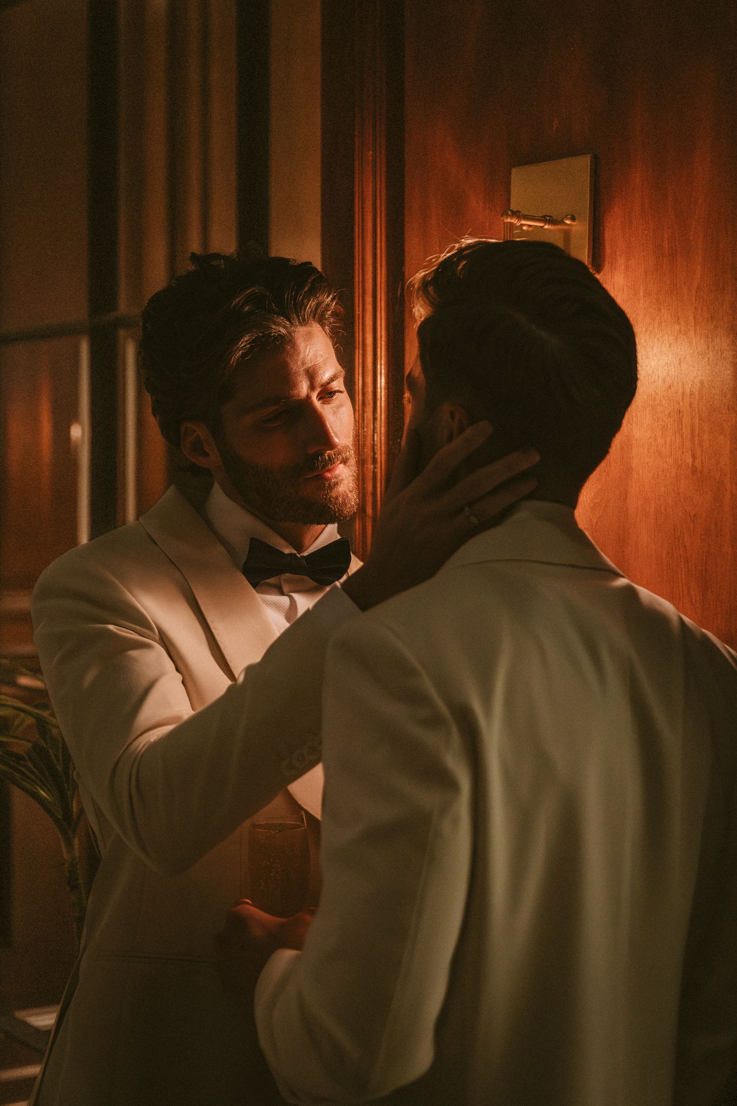 Two grooms in white tuxedos tenderly embracing in a warmly lit corridor before wedding ceremony.
