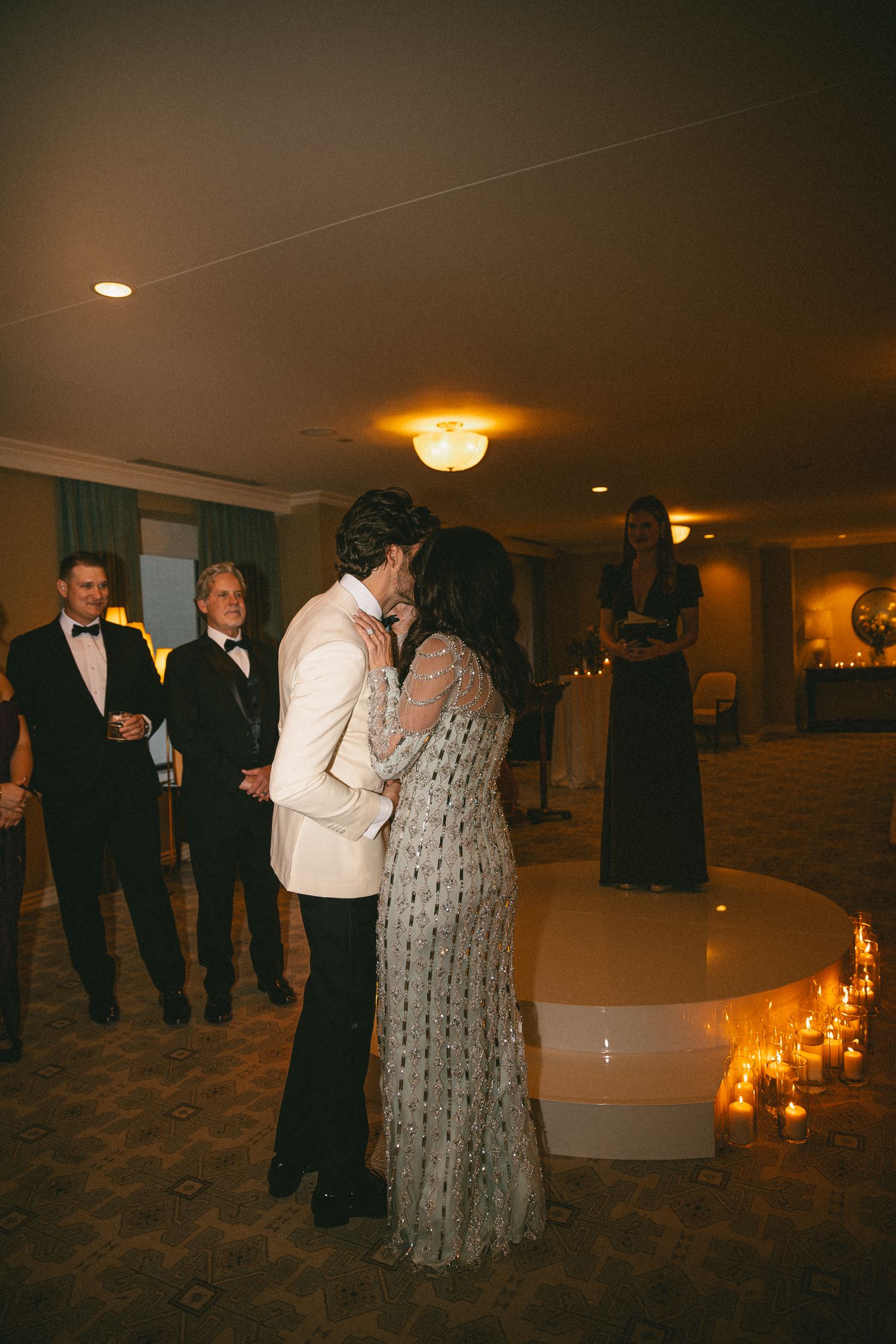 Couple sharing first dance kiss in dimly lit wedding reception hall surrounded by candlelight and guests.