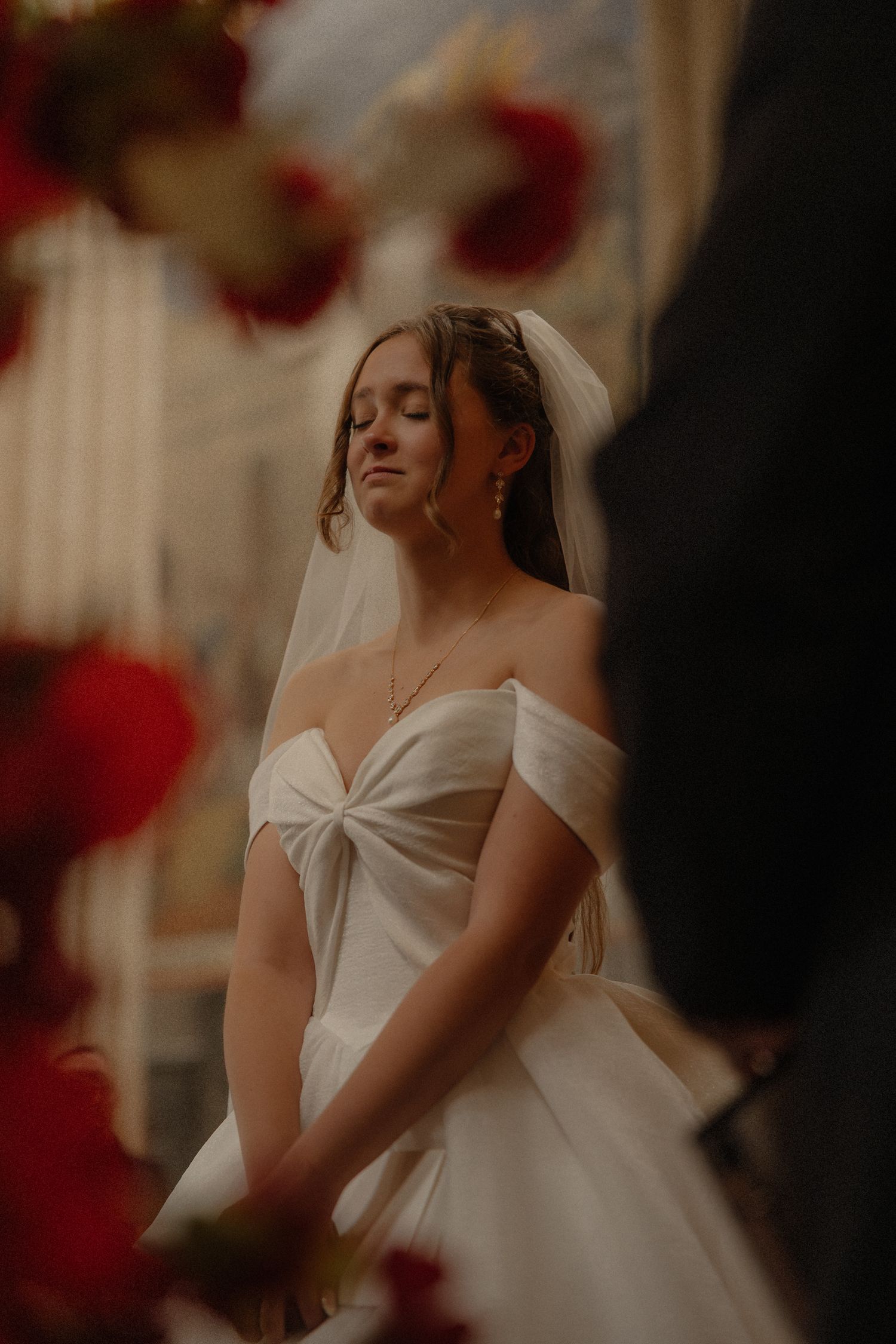 Bride in off-shoulder white wedding dress with eyes closed under red floral arch.