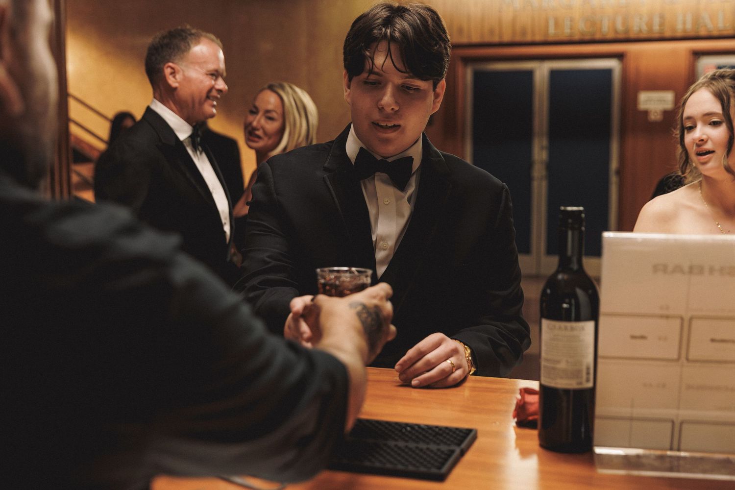 Groom in tuxedo holding drink at bar surrounded by wedding guests in elegant venue.