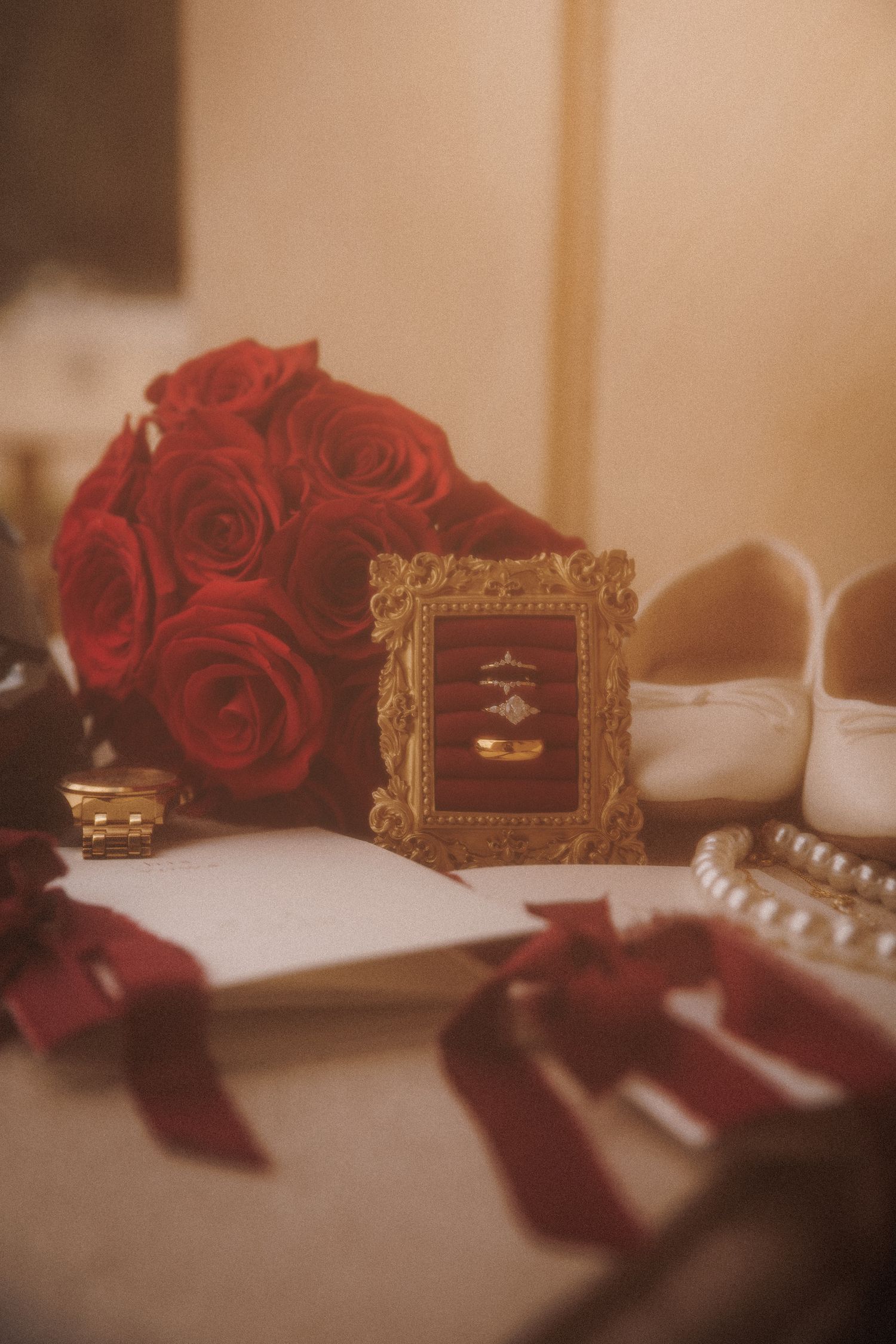 Elegant wedding flat lay with red roses, gold rings in ornate frame, open journal, pearl bracelet, and ribbon accents.
