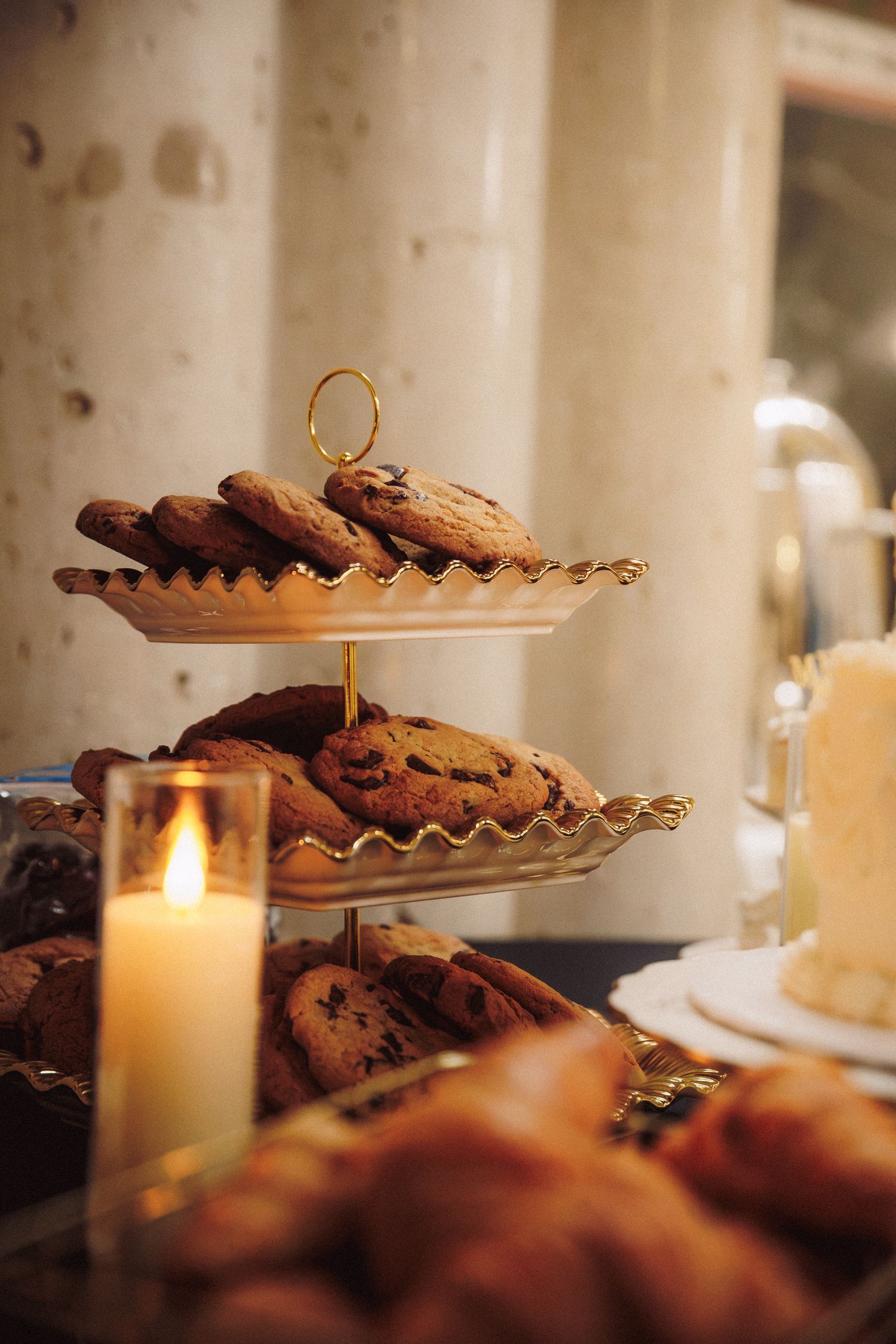 Gold tiered tray with chocolate chip cookies and pastries beside a lit candle at an elegant wedding reception.