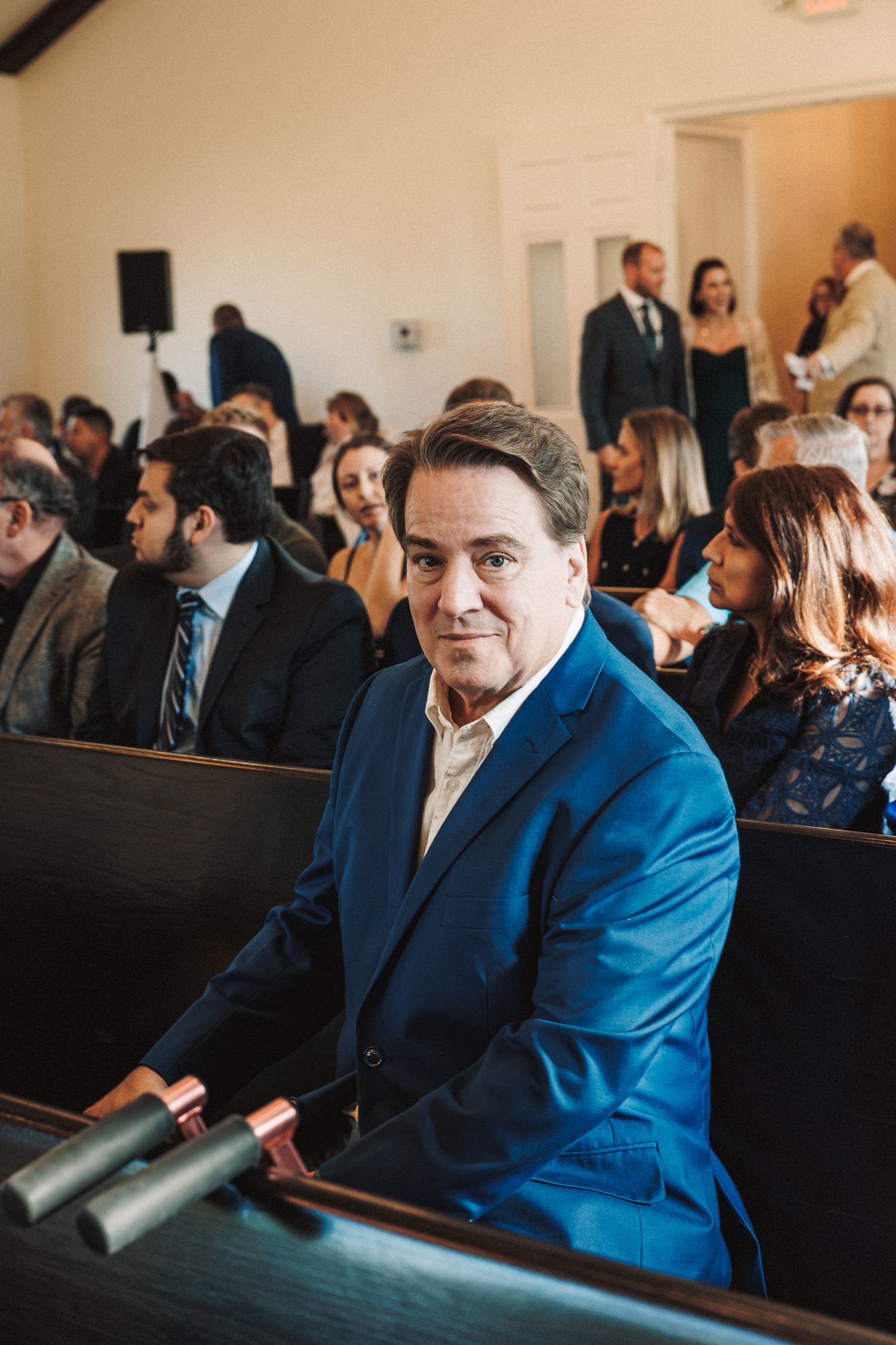 Wedding guest in blue suit smiling at camera while seated in church pew.