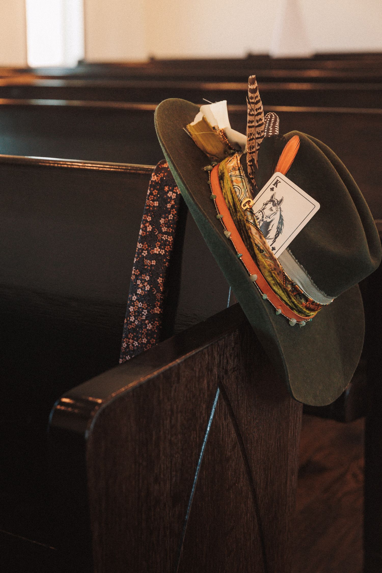 Decorative cowboy hat with feathers and playing card resting on dark wooden church pew.