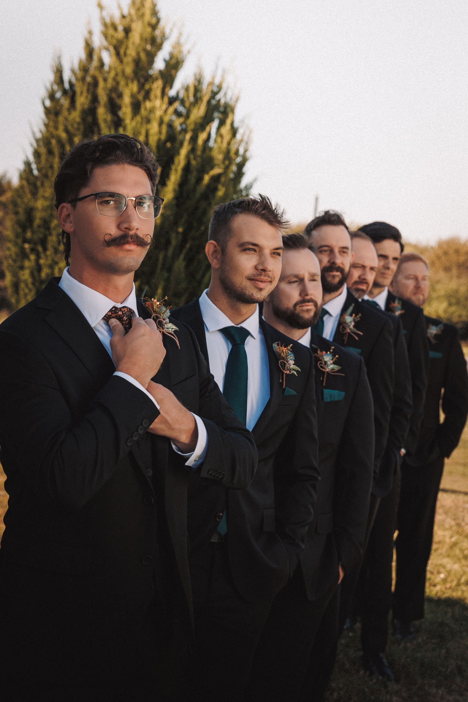 Groomsmen in black suits with boutonnieres lined up outdoors at golden hour wedding.