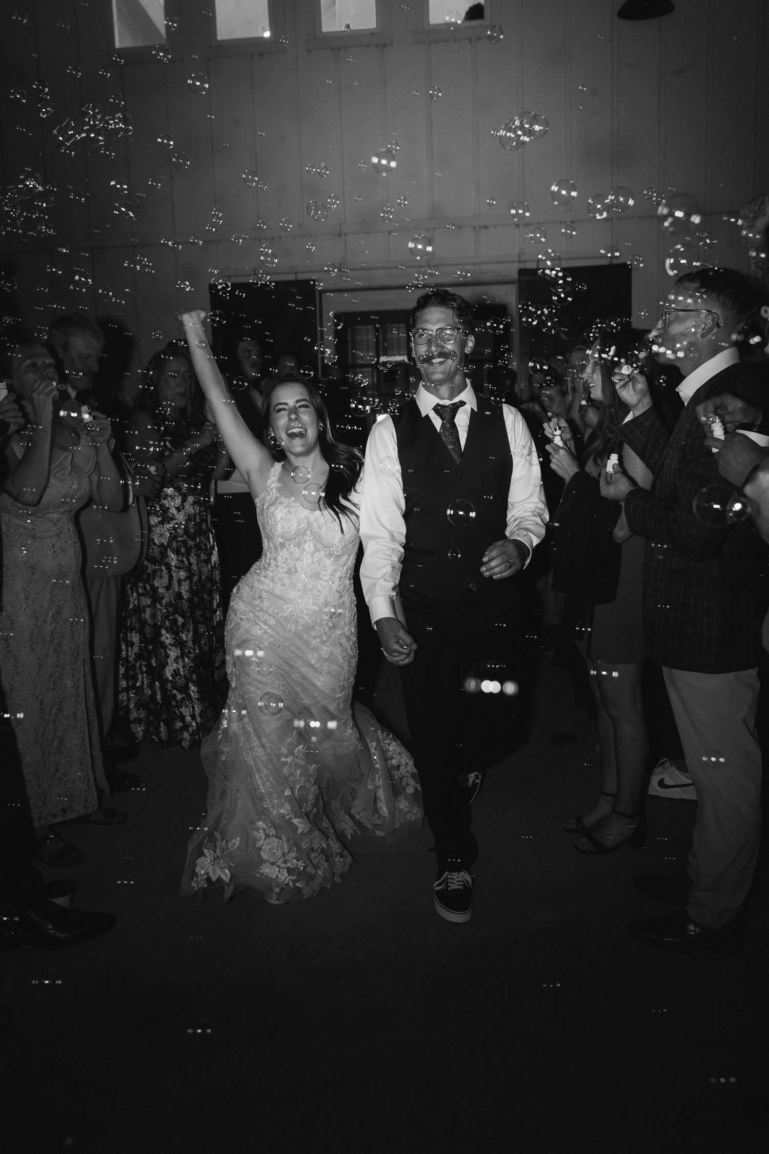 Bride and groom dancing joyfully surrounded by bubbles and guests in black and white wedding photo.
