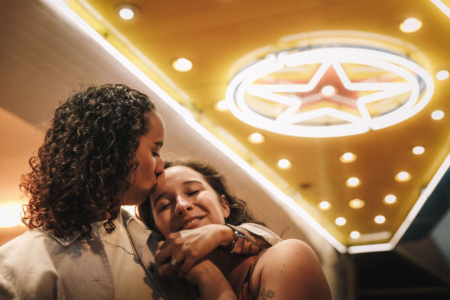 Two women share a tender moment under glowing star neon lights in a warmly lit venue.