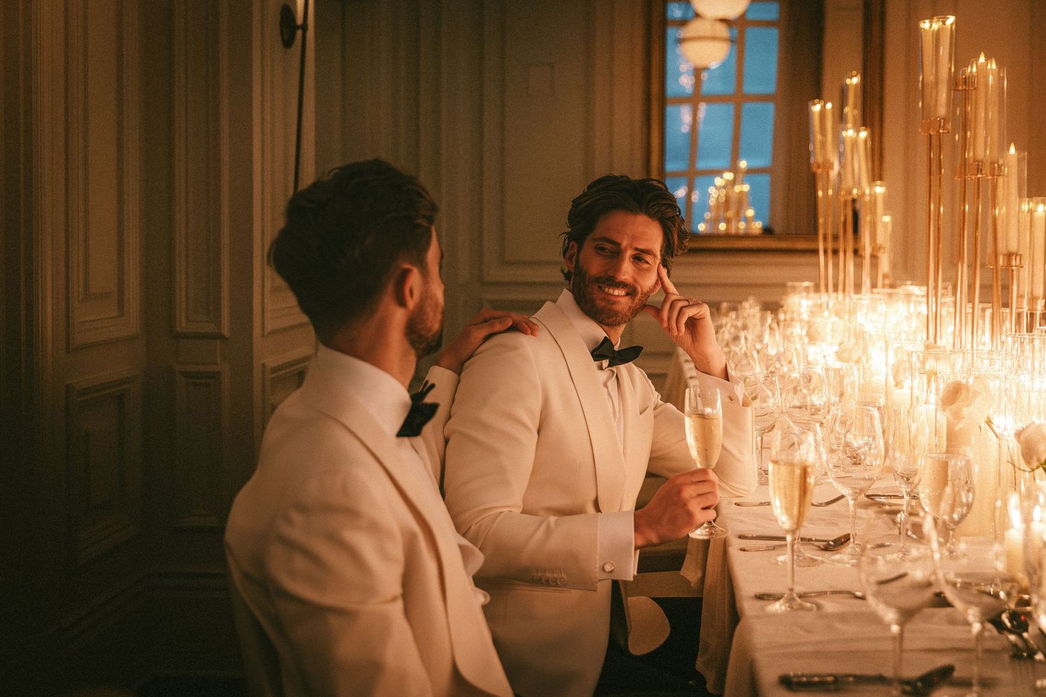 Two grooms in white tuxedos laugh together at a candlelit wedding reception dinner table.