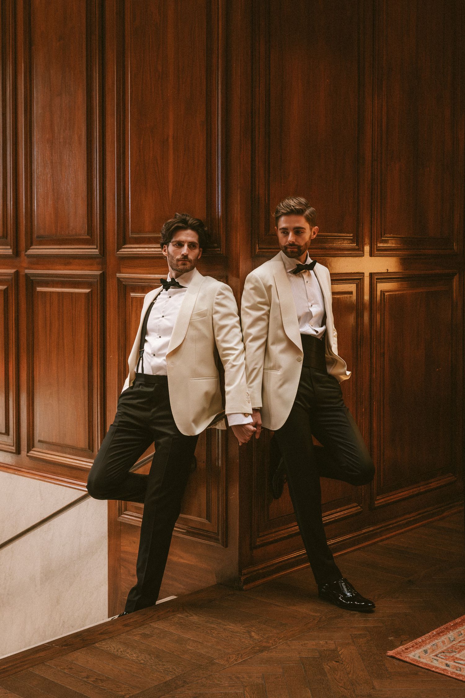 Two grooms in matching ivory tuxedos hold hands against ornate wooden paneled walls.