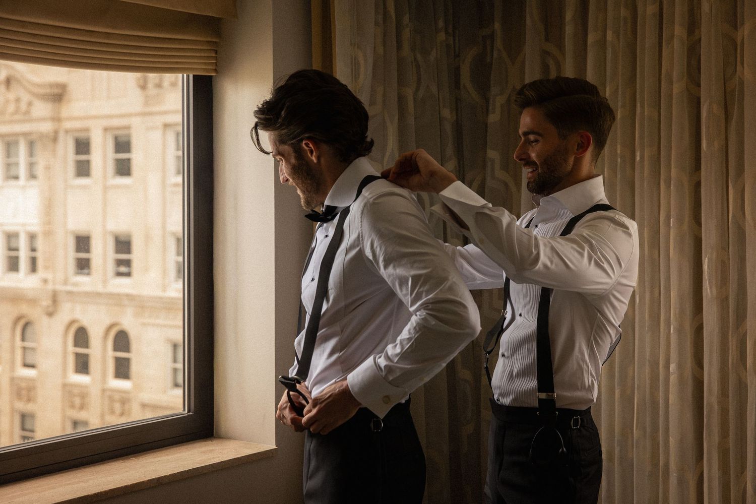 Two grooms in white shirts and suspenders help each other get dressed near a sunlit window before wedding.