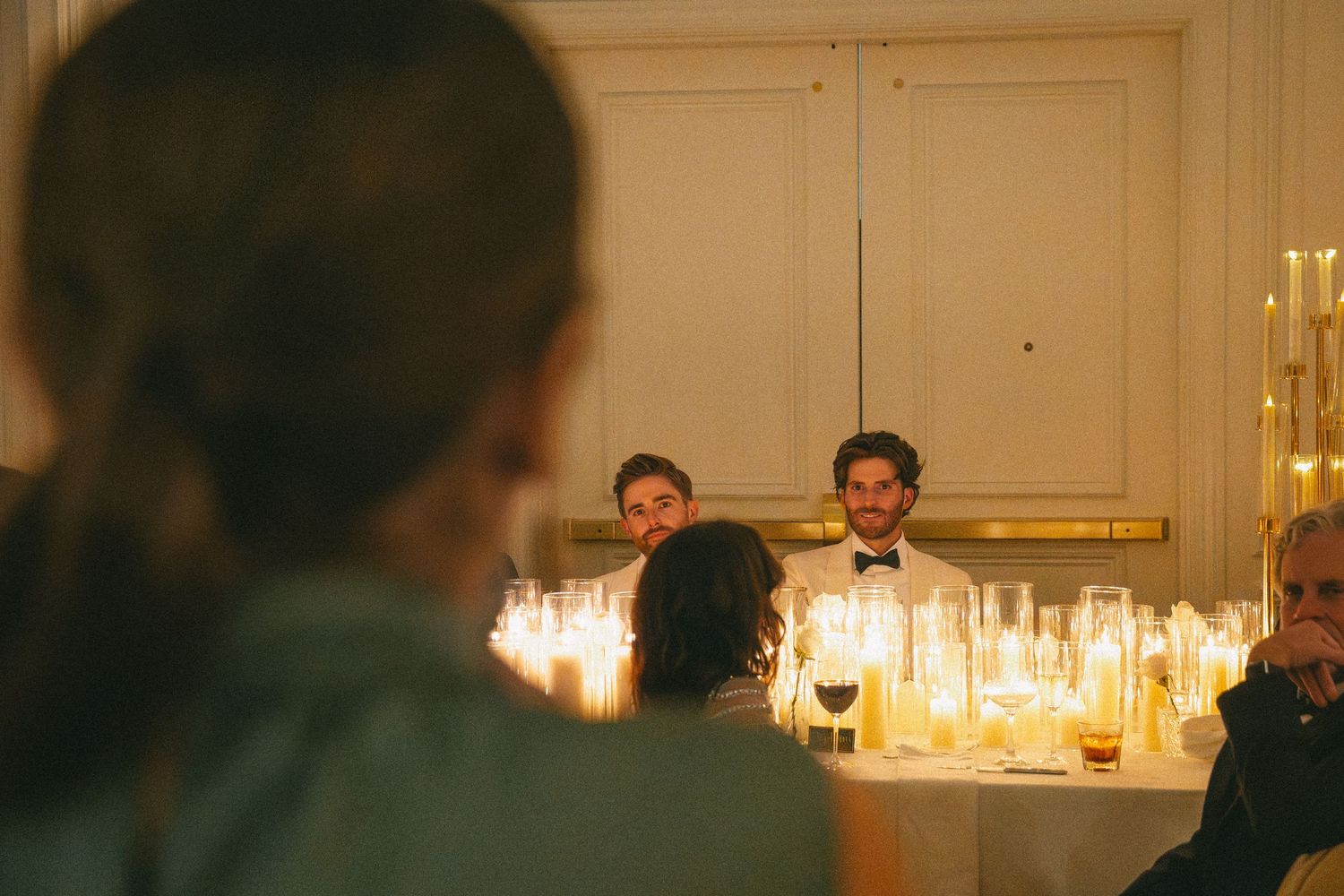 Two grooms smiling at their candlelit sweetheart table during an intimate wedding reception.