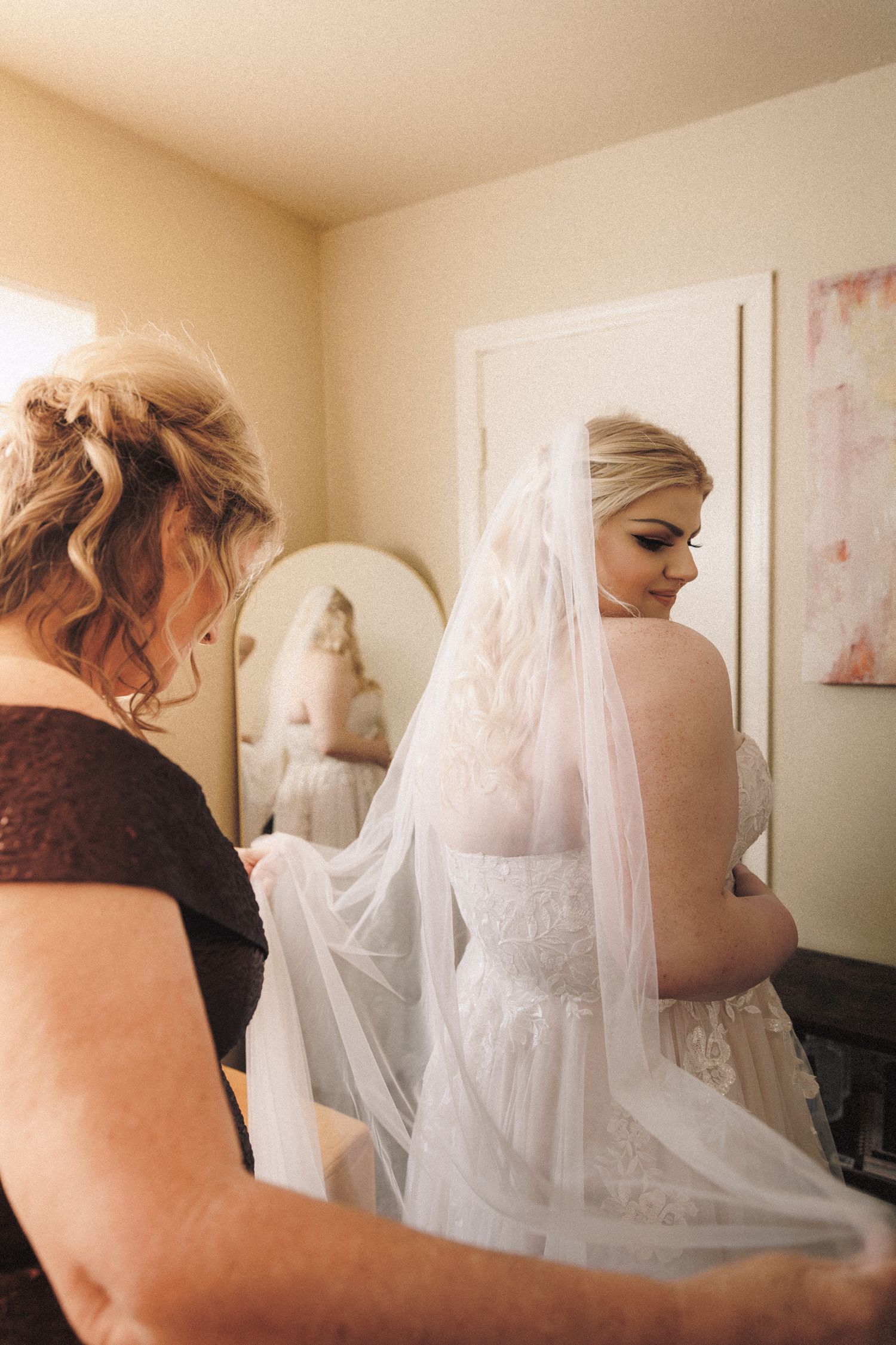 Mother helping bride with flowing white veil and lace wedding dress before ceremony.
