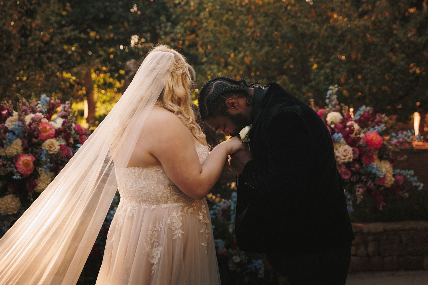 Bride and groom share first kiss at altar surrounded by colorful floral arrangements outdoors.