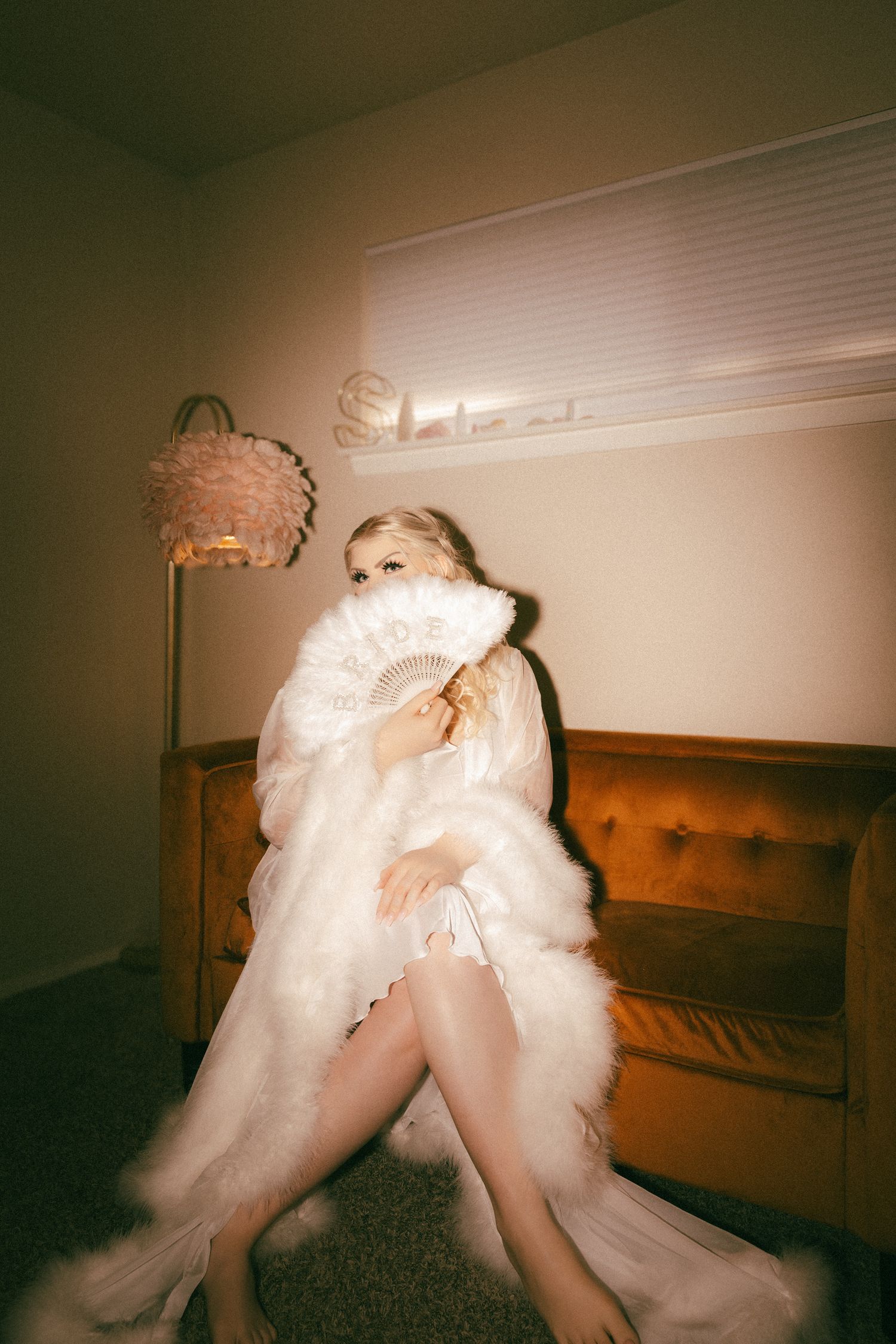 Blonde bride in white fur robe holding feather fan on retro velvet sofa in warm lit room.