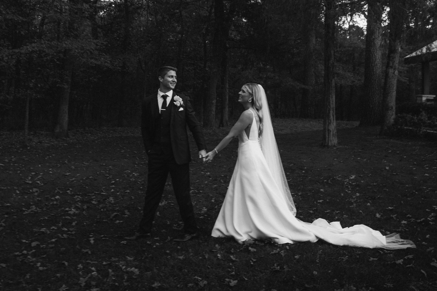 Black and white photo of bride and groom holding hands in wooded forest setting.