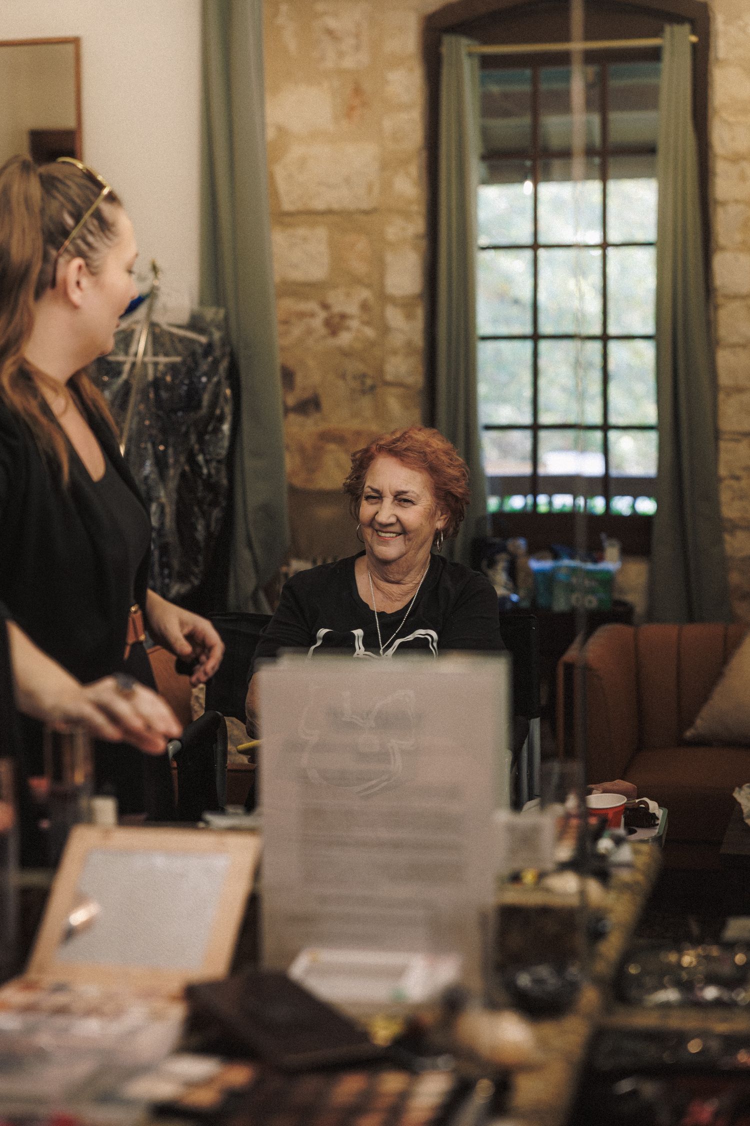 A smiling woman with red hair laughs during wedding preparations at a makeup and styling station.
