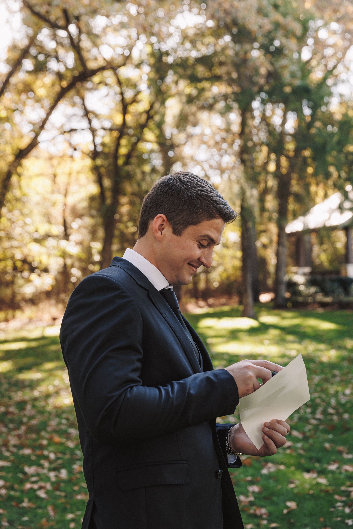 Handsome groom in dark suit smiling while reading a letter outdoors in an autumn garden setting.