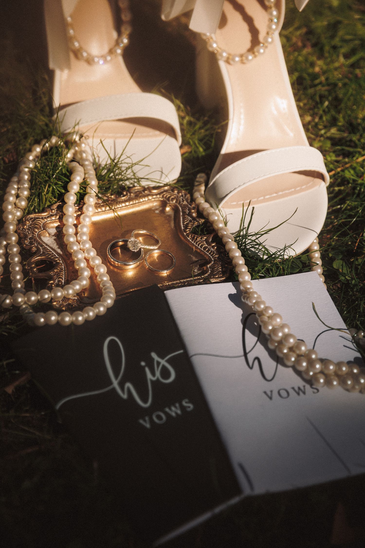 Wedding flat lay with white heels, pearl necklace, gold rings, and his and hers vow books on green grass.