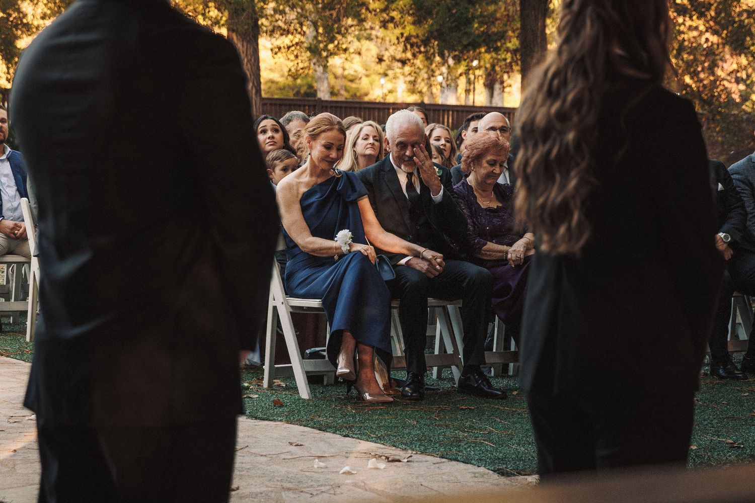 Wedding guests seated outdoors watching a ceremony, with a woman in blue dress smiling in the front row.